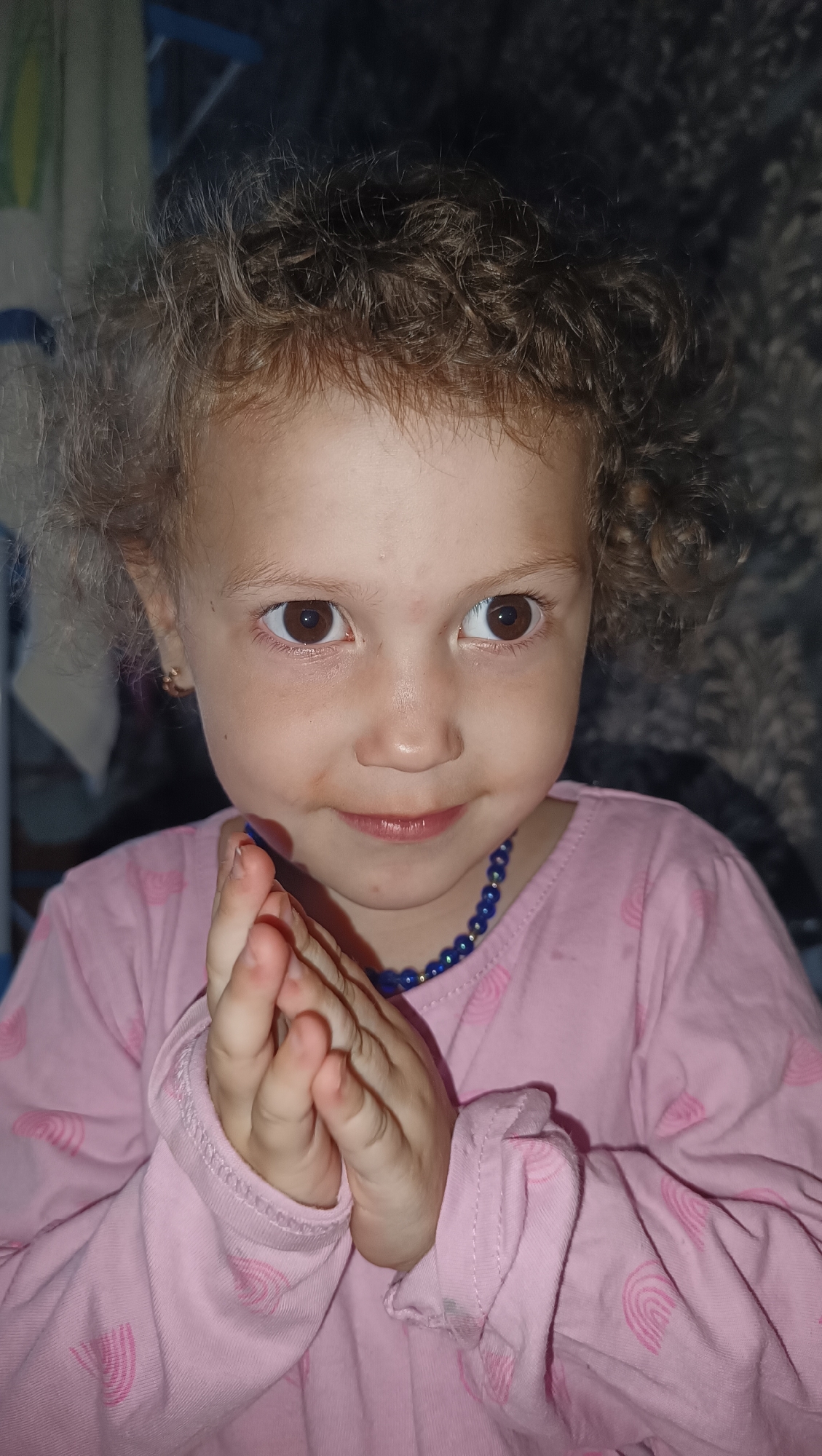The image is a close-up portrait of a young girl with curly brown hair. She is wearing a pink long-sleeved shirt with a pattern of pink rainbows. She has a blue beaded necklace around her neck. Her hands are clasped together in front of her chest, and she is looking at the camera with a slight smile. Her eyes are brown, and she has rosy cheeks. The background is blurred and appears to be a dark, textured surface. There is no discernible information about the city of Karsha, Russia, or any other specific location. The image appears to be a candid photograph taken indoors, likely in a domestic setting.