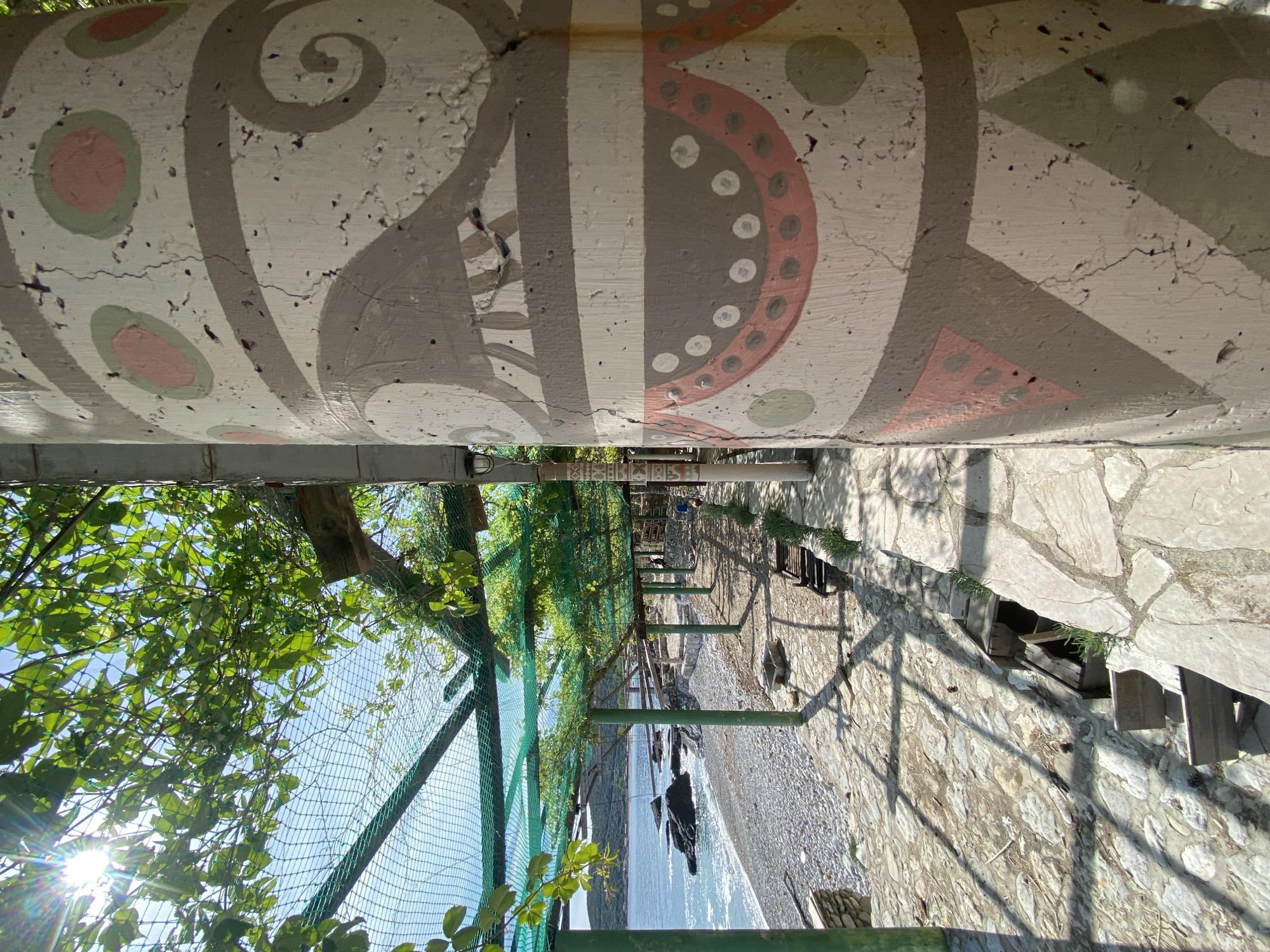 The image depicts a view from underneath a painted concrete structure in Prijevor, Montenegro. The structure features a repeating pattern of stylized, circular, and triangular designs in white, gray, and muted pink. Below the structure, a metal-framed fence runs along a path towards the sea. Lush greenery grows on the left side of the fence. The pathway, constructed from irregular stone slabs, leads to a pebbled beach and the sea, which is visible in the distance. Shadows from the fence and overhead structures are cast on the pathway and beach. The sun is visible through the leaves.