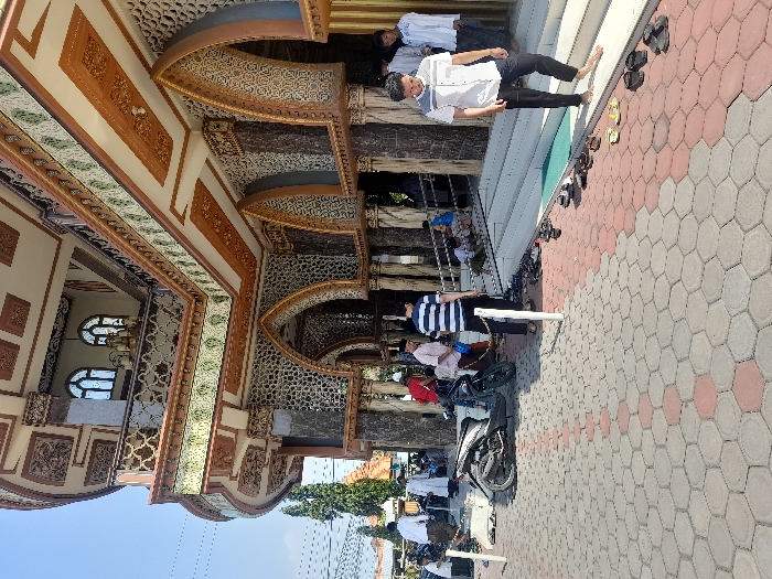 The image shows the exterior of a mosque in Kecamatan Candi, Indonesia, located at latitude: -7.494, longitude: 112.717. The mosque has an intricate facade with intricate details, including arches, columns, and decorative tile work. There are people standing in front of the mosque. Some are wearing traditional Muslim clothing, while others are wearing casual attire. A black motorcycle is parked in front of the mosque. The people are interacting with each other and appear to be waiting to enter the mosque. There are shoes and sandals lined up on the steps leading up to the mosque. The environment is sunny and bright.
