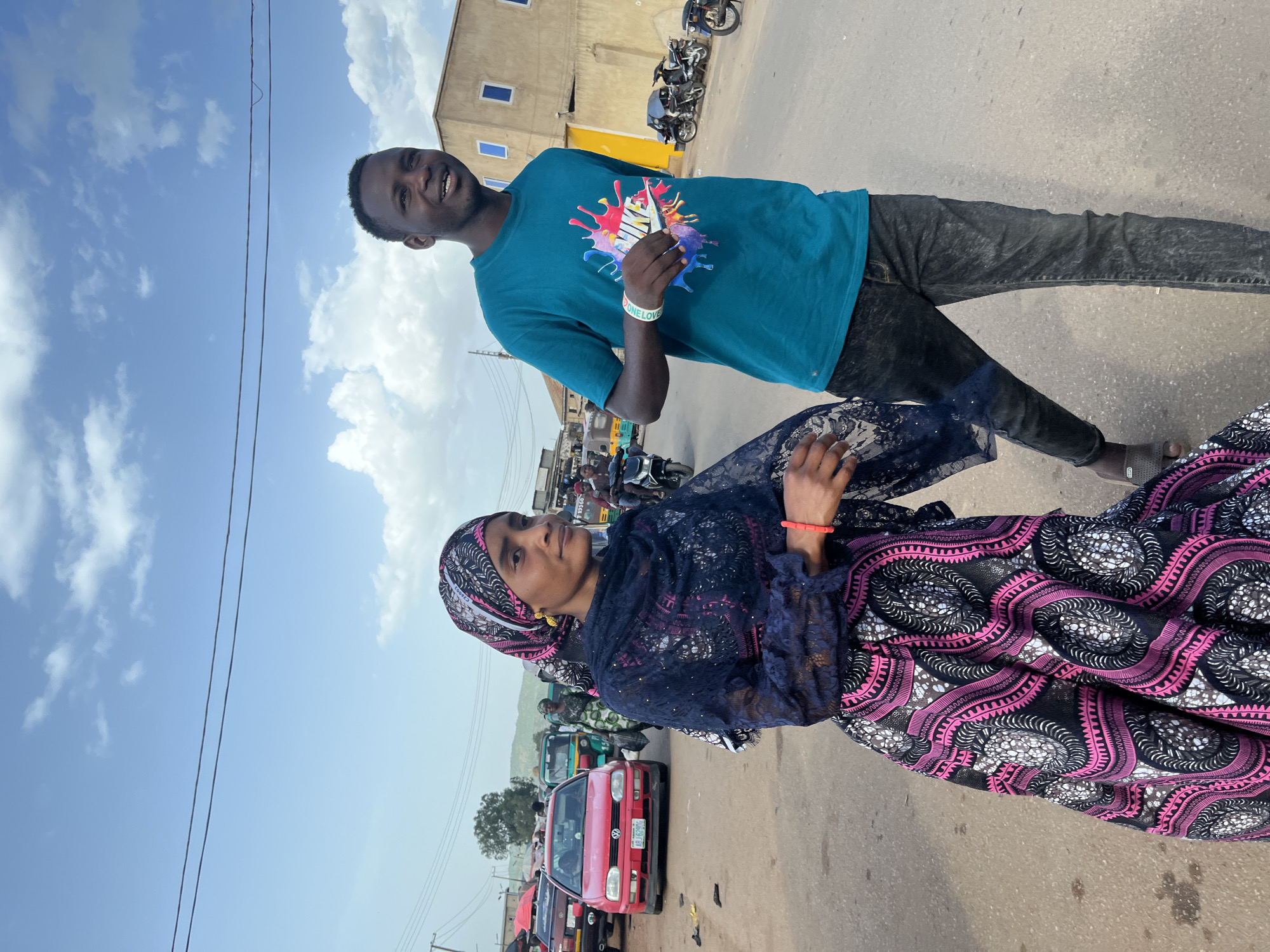 In Rafin Par, Nigeria, a man and a woman walk side by side on a dusty, paved road. The woman, wearing a black and pink patterned dress and headscarf, looks slightly away from the camera, her right arm adorned with a red bracelet. The man, in a teal t-shirt with a colorful paint-splatter design and dark jeans, faces the camera and smiles. He has a white wristband on his left arm. In the background, a red Volkswagen car, a green auto-rickshaw, and several motorcycles are visible, along with a two-story building with yellow and light-colored walls and blue-framed windows. The sky is a vibrant blue with scattered white clouds.