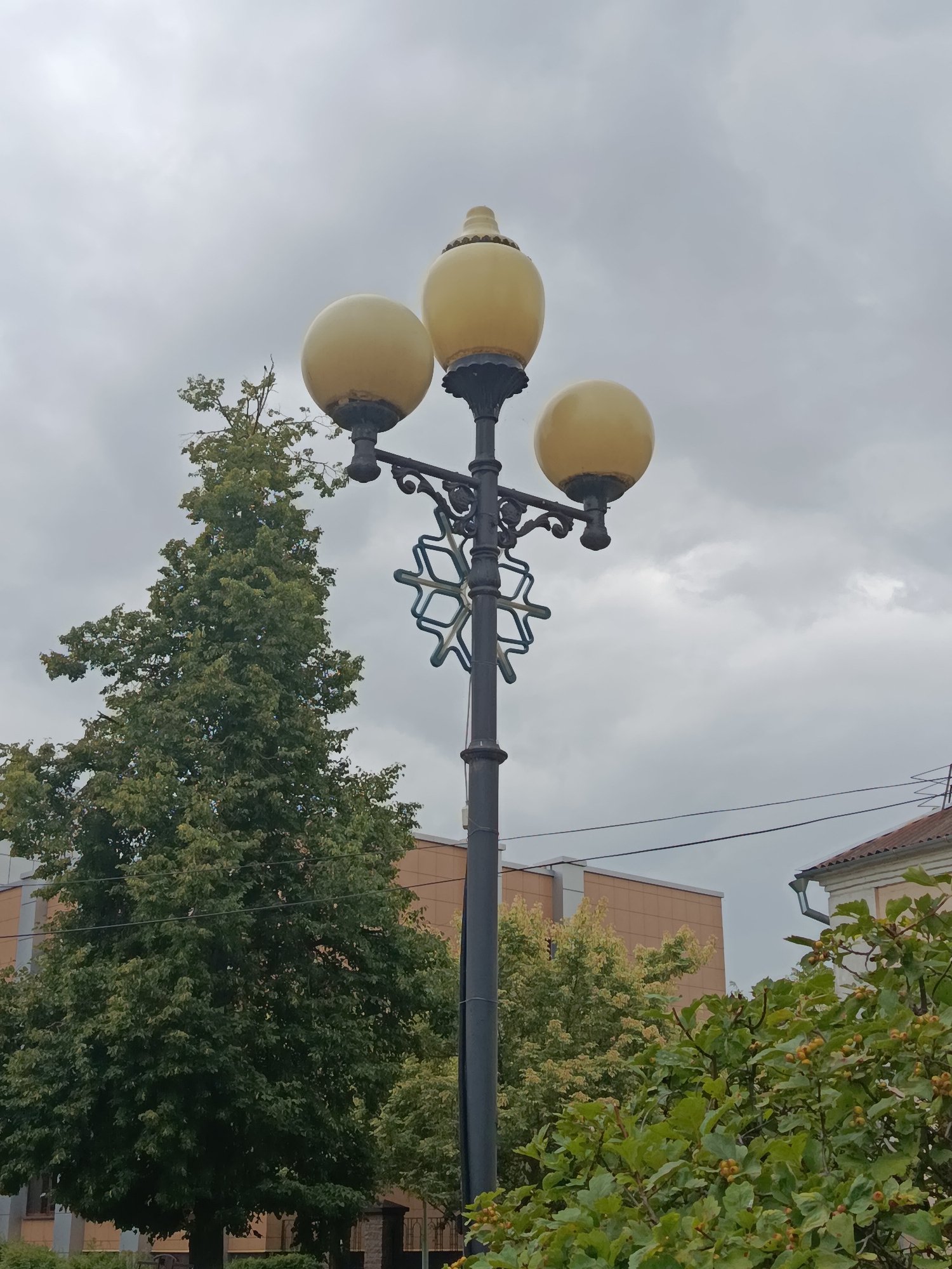 The image depicts a street lamp with three yellow, globe-shaped lights against a cloudy sky. The lamp post is black and ornate, featuring a decorative snowflake-like metal accent. To the left of the lamp post is a large, full green tree. In the background, there are modern buildings with beige and cream-colored facades, and some foliage. The overall scene appears to be an outdoor urban environment. There are no people or animals visible in the image. The image is taken from a low angle, looking upwards at the lamp post and trees.