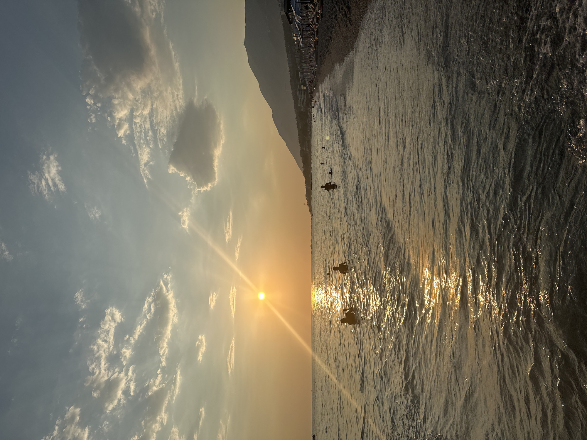An aerial drone shot captures a tranquil beach scene at sunset in an unspecified location. The sun, a bright orb low in the sky, casts a golden glow across the tranquil sea, creating a shimmering path of light on the water's surface. Several silhouettes of people are visible wading in the shallow waters, enjoying the warm evening. The shoreline is lined with beach chairs and umbrellas, hinting at a populated tourist destination. Majestic mountains rise in the background, their peaks silhouetted against the soft hues of the sky, where scattered clouds drift lazily. The overall atmosphere is one of peace and relaxation, characteristic of a perfect summer evening at the coast.