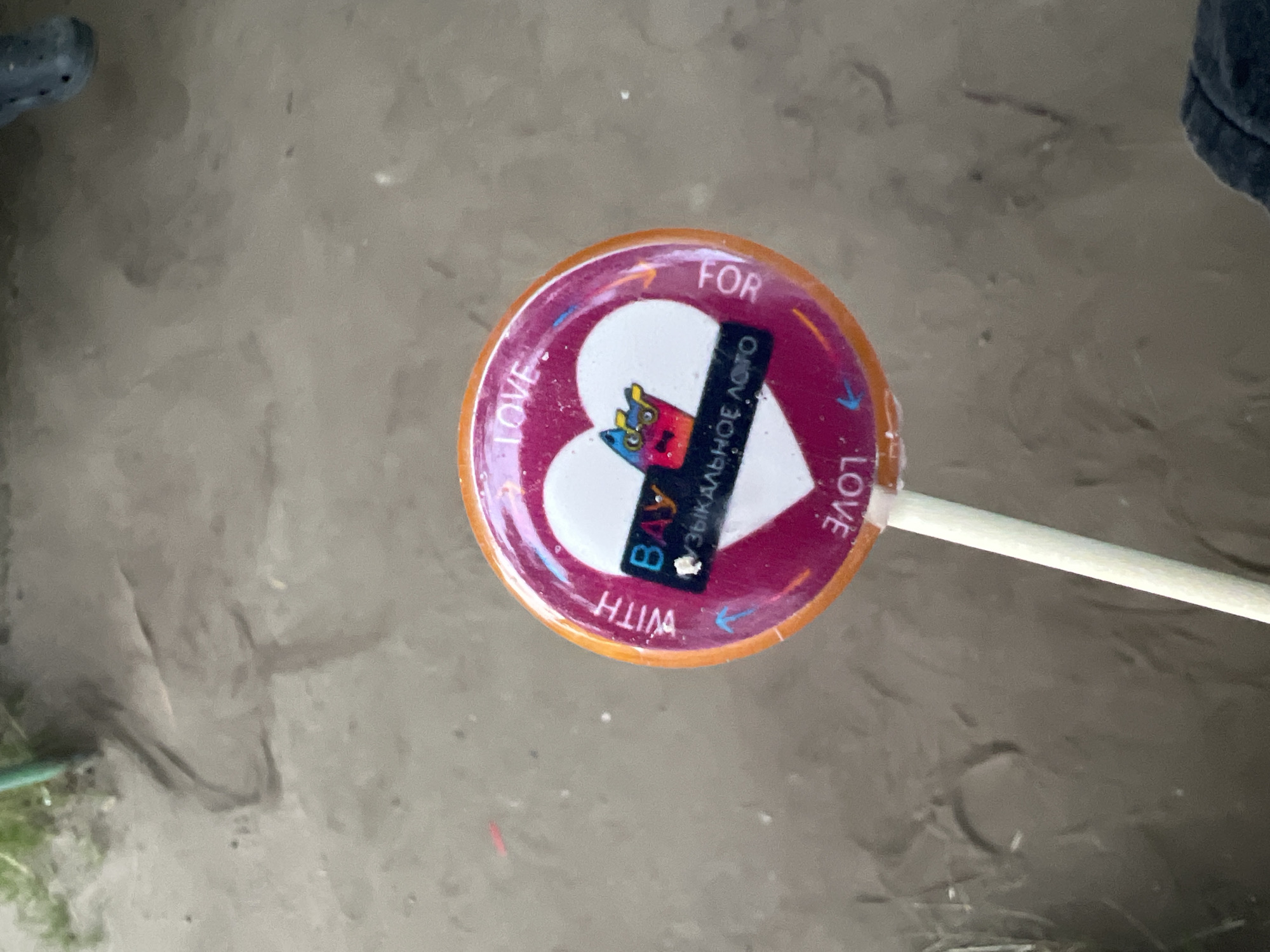 A lollipop with a heart-shaped design is held up by a white stick. The lollipop has an orange border. Inside the heart, there is a cartoon image with the words "BAY" and "МУЗЫКАЛЬНОЕ ЛОТО" (Musical Lottery) in Russian. The words "WITH LOVE FOR" are written around the edge of the lollipop in English, with some arrows indicating direction. The background appears to be sandy ground, possibly a park or outdoor area, with some dried grass and possibly debris. The image is taken from a slightly elevated perspective, looking down at the lollipop. There are no discernible landmarks to pinpoint a specific location in Ulyanovsk, Russia.
