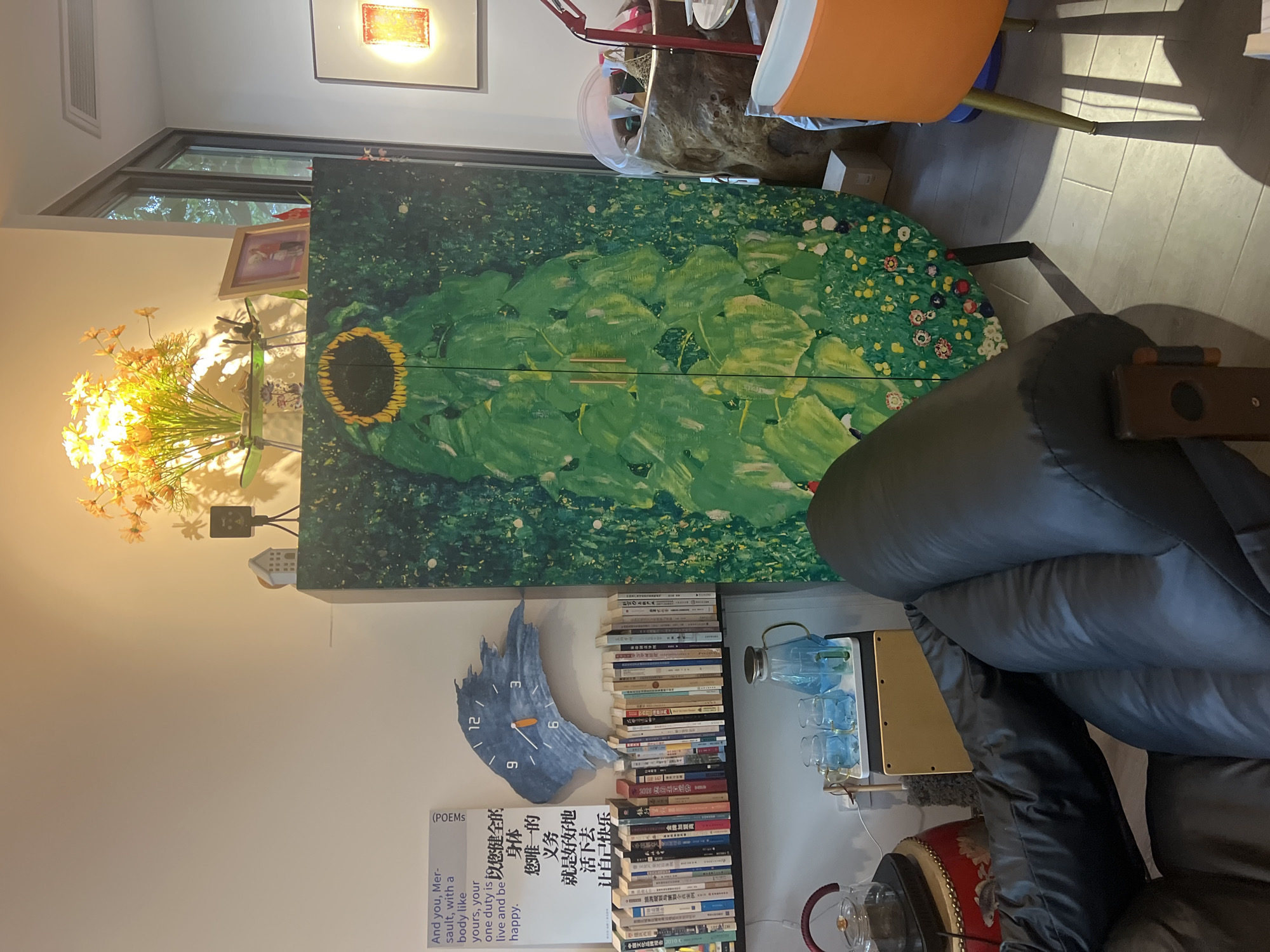 The image depicts an indoor scene, possibly a living room or study, with furniture and decorative items. The focal point is a tall, green cabinet featuring a large, stylized sunflower painting. To the left of the cabinet, a bookshelf is filled with books. Above the bookshelf, a blue, irregularly shaped clock is mounted on the wall. A framed picture and a bouquet of orange flowers with a spotlight are visible on top of the cabinet. In the foreground, a black leather sofa is partially visible. In the background, an orange chair and a large, textured urn-like object are present. The lighting suggests a warm, ambient environment. The presence of Chinese characters on some of the books and posters indicates a likely location in China, potentially within Liandu District. There are no people visible in the image.