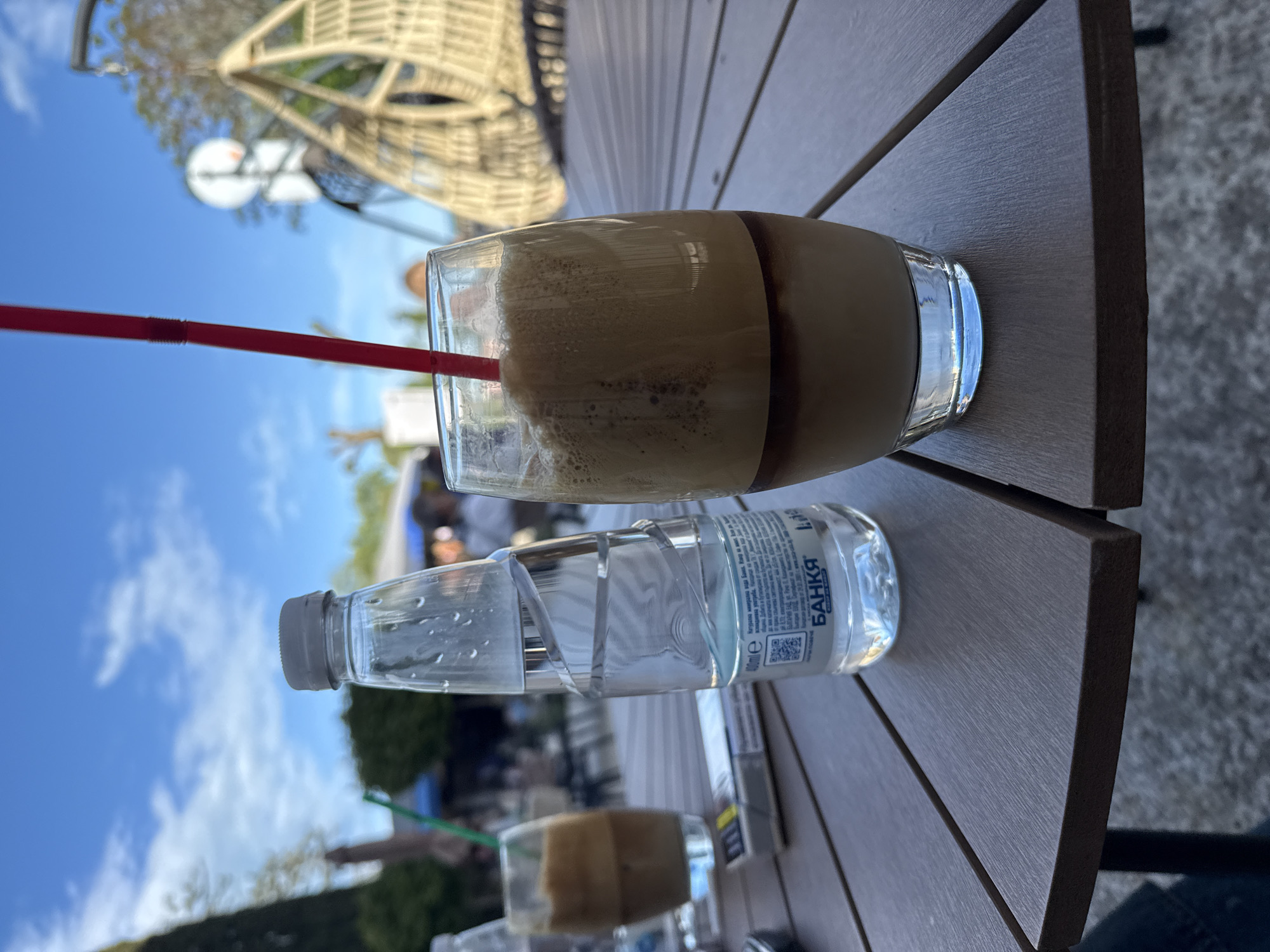 The image shows a table with a glass of coffee, a water bottle, and a red straw. The bottle has a label with "БАНКА" (Bankia) on it. In the background, a structure and blue sky are visible. The image appears to be taken at a cafe in Varna, Bulgaria. Another person is present in the frame.