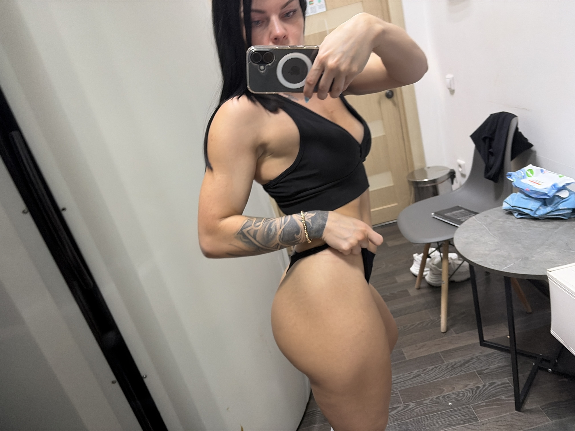 The image shows a woman taking a mirror selfie in what appears to be a dressing room or a private space with light-colored walls and wooden floors. She is wearing a black sports bra and black thong, showcasing her muscular physique and tattoos on her left arm. She is holding a smartphone in her right hand, with the camera facing a mirror.
In the background, a gray chair is visible with a black garment draped over it, and a round, gray table is set up with a stack of blue packaged items. To the left of the woman, a dark vertical structure, possibly a door or partition, is present. There is a metallic trash can near a wooden door in the background. The lighting suggests it could be daytime, but no specific time is discernible. The overall mood is casual and self-observational. No visible text is present in the image. The location context provided is Ufa, Russia.