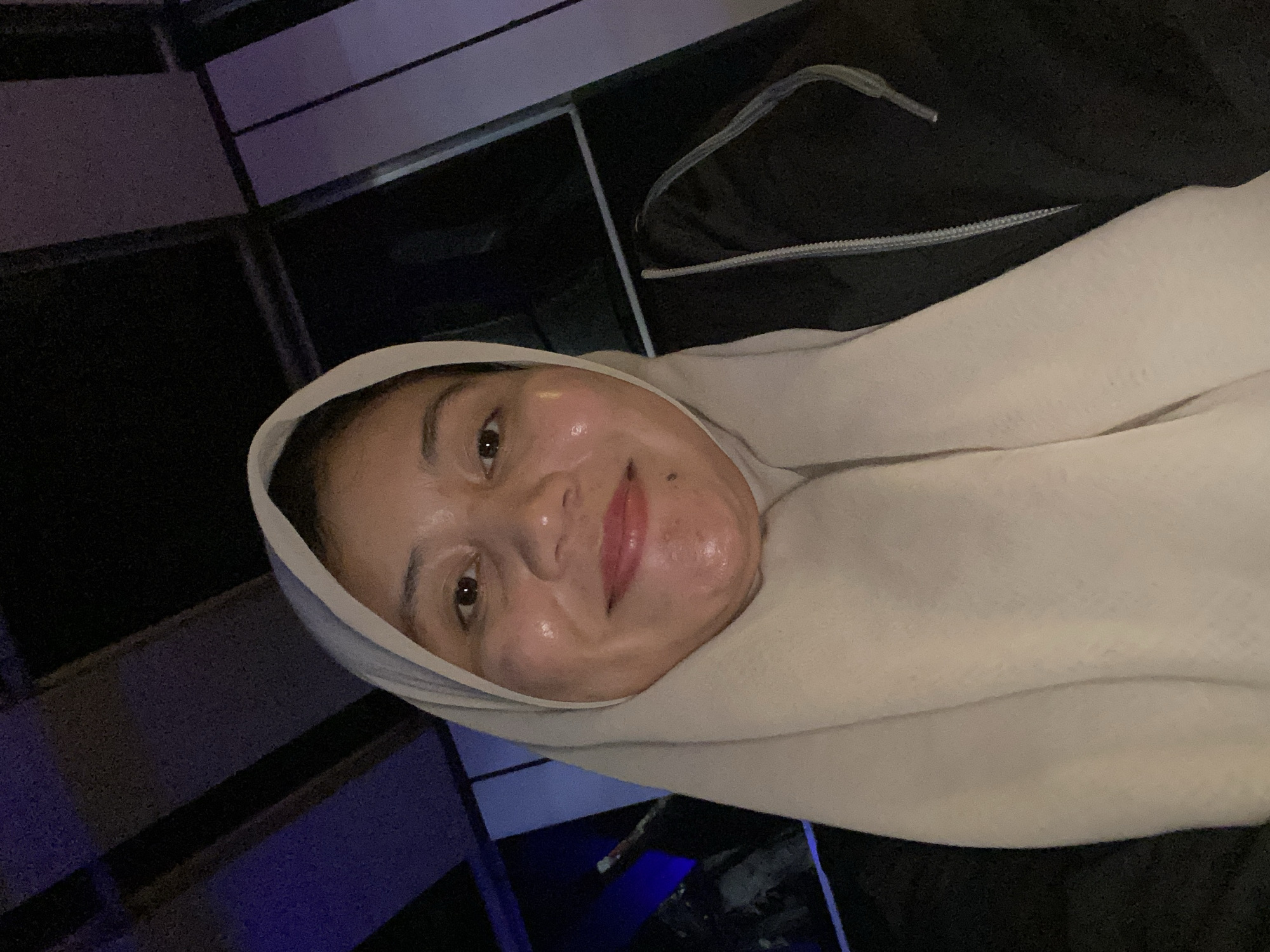 This is a close-up, self-portrait of a young woman wearing a light-colored hijab. Her face is turned towards the camera, and she is smiling. She appears to have a dewy complexion and is wearing light pink lipstick. She is wearing a dark-colored jacket, and the hijab is a pale beige color. The background is dark and out of focus, but it appears to be an indoor setting with some dark, geometric shapes and blue lighting, possibly indicative of a modern interior or a dimly lit public space. The image is oriented sideways. The time of day and weather are not discernible from the image. There is no visible text. The location context of Banjarbaru, Indonesia is provided.