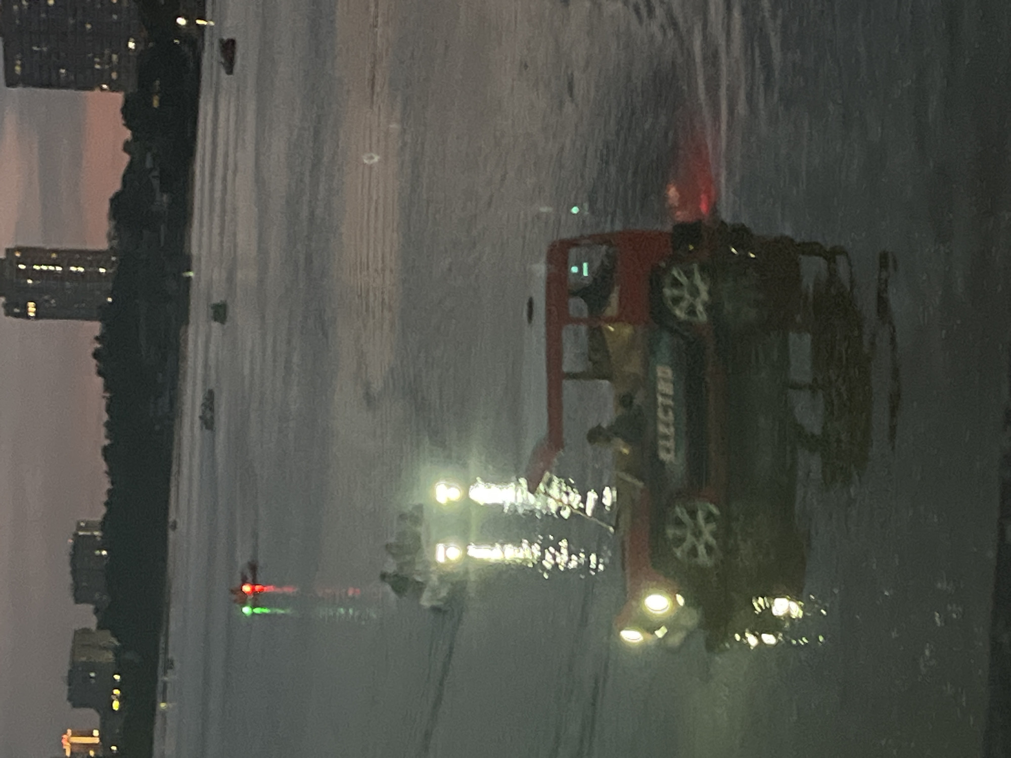 The image shows a body of water with several boats, some of which appear to be amphibious vehicles designed for both land and water. In the foreground, a red amphibious vehicle with white headlights is partially submerged, moving through the water. The word "ELECTRO" is visible on its side. In the middle ground, another boat with people on board is illuminated by its headlights. Further in the background, a smaller boat with green and red lights is visible. The scene is set at dusk, with the skyline of a city featuring tall buildings in the distance. The water is calm, reflecting the lights from the vehicles and the city. The overall impression is of a recreational or transportation activity taking place on the water at twilight.