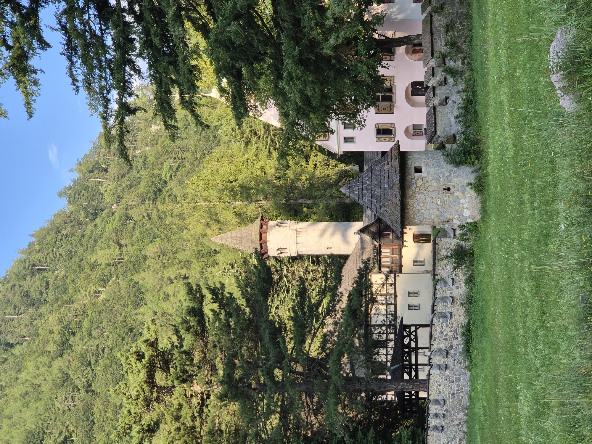 This image captures a portion of the historic Erzberg mountain village in Eisenerz, Austria, with the iconic "Small Fortress" (Kleiner Bergfried) as a prominent feature. A tall, stone tower with a conical roof, resembling a watchtower or small keep, stands to the left, exhibiting weathered stone masonry and small rectangular windows. Adjacent to it, a section of a building with a light-colored facade and dark timber framing is visible, suggesting a traditional Austrian architectural style. To the right, a pastel pink building with arched windows and wooden shutters adds a touch of color to the scene.
A stone wall, constructed from irregularly shaped rocks and topped with wooden crenellations, encloses the area, evoking a sense of historical fortification. In front of the wall, a small, round stone turret with a shingled conical roof provides a charming contrast to the larger tower. The foreground is dominated by a lush green meadow, dotted with wildflowers and tall grasses, indicative of a natural, outdoor setting.
Towering behind the structures, a dense forest covers the steep slopes of the Erzberg mountain, with the bright blue sky visible above. The sunlight casts shadows, highlighting the textures of the stone, wood, and foliage, and suggesting it is either early morning or late afternoon. No people or animals are visible in the frame. The overall impression is one of a serene, historically significant landscape, nestled within the natural beauty of the Austrian Alps.