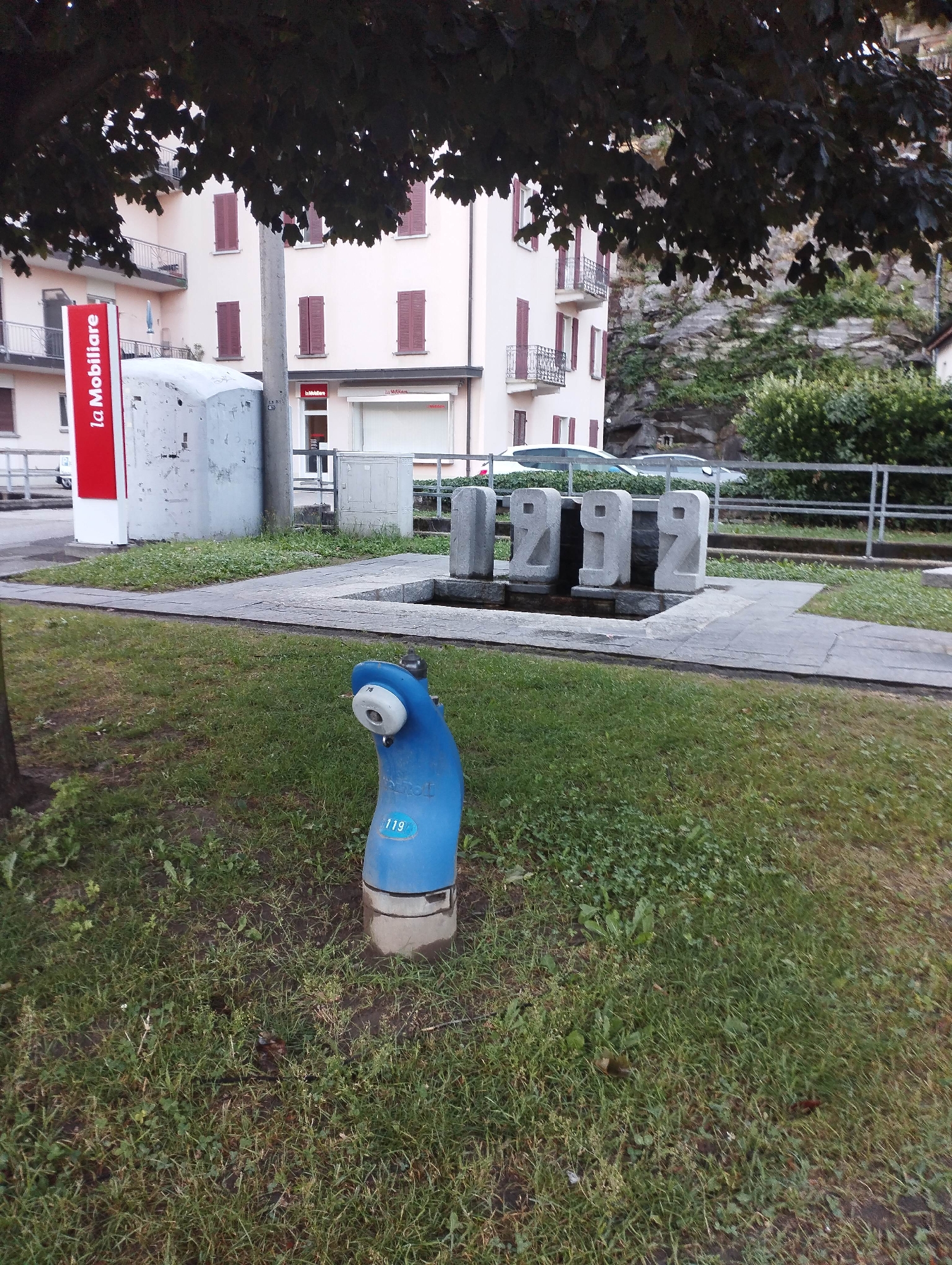 The image captures a static outdoor scene, likely a public space or park area, on what appears to be an overcast day. In the foreground, a vibrant blue, irregularly shaped object, possibly a water dispenser or public fountain, stands on a patch of green grass. It has a light-colored spout or sensor on its curved top, and the number "119" is visible on its side, along with partially obscured text that seems to include "totem." The ground is covered in grass, with a small tree trunk visible to the left.
In the midground, a paved pedestrian path separates the grassy area from a rectangular stone structure featuring the large, carved numbers "1932." This structure appears to be a monument or part of a small fountain. Behind it, a metal railing runs along what looks like a drop or a slight incline, beyond which two cars are parked.
The background features a light-colored, multi-story building with numerous windows fitted with closed red shutters and balconies with metal railings. A prominent red sign reading "La Mobiliare" is visible next to this building, suggesting a commercial or residential complex. Another smaller "La Mobiliare" sign is seen on a shopfront. Further back and to the right, a natural rock face covered with some greenery rises steeply, hinting at a mountainous or hilly environment.
No people or explicit activities are taking place; the scene is quiet and still. The time of day is likely daytime, with soft, diffused light indicating either an overcast sky or possibly early morning/late afternoon. The "La Mobiliare" text, the architectural style with red shutters, and the "1932" monument could suggest a European location, possibly Italian or Swiss.
