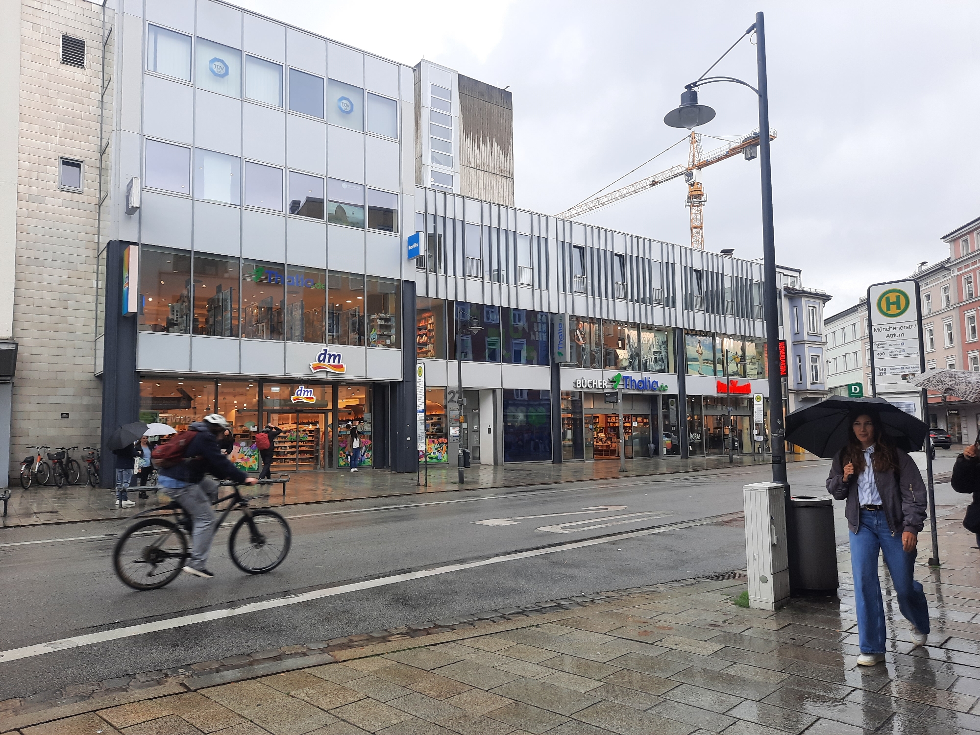 The image depicts a street scene in Rosenheim, Germany, on a cloudy, wet day. In the foreground, a person on a bicycle is in motion, their form blurred due to the movement. To the right, a woman walks under a black umbrella, dressed in a grey jacket and blue jeans, with other pedestrians visible in the background also using umbrellas.
The street itself is wet and reflective, indicating recent rain. A bus stop sign for "Münchener Str./Atrium" is visible on the right side of the image, identifying the location. Adjacent to the street are shopfronts, including a "dm" drugstore and a "BUCHER Thalia" bookstore, with their interior lights on. A construction crane stands prominently in the background, suggesting ongoing building work in the area. Bicycles are parked along the sidewalk on the left side of the street. The buildings lining the street are a mix of modern architectural styles.
