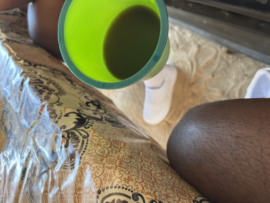 A green plastic cup filled with dark liquid sits above a patterned surface covered in clear plastic. Partially visible are two legs, one fair-skinned with a white sock and the other with dark skin. A metallic surface appears in the top right corner. The scene is captured from an overhead perspective.