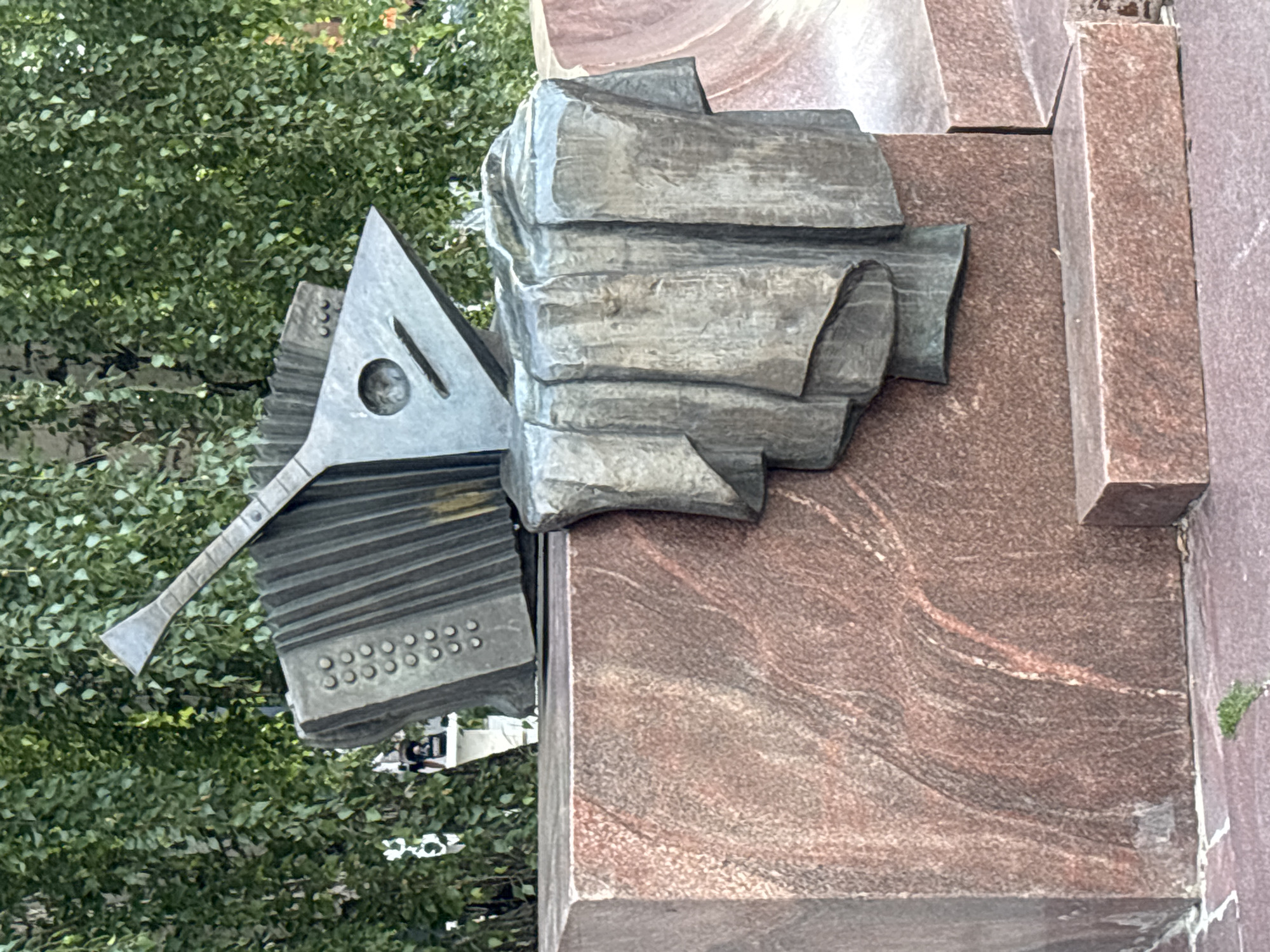 A bronze sculpture of an accordion and a balalaika rests atop a pink granite monument in Novaya Usman', Russia. The accordion, depicted in an open state, is positioned to the left, while the balalaika, a triangular stringed instrument, is placed on top of it. A draped cloth-like element, also made of bronze, cascades from the accordion and balalaika onto the granite base. The monument's base is a large, rectangular block of pink granite with visible veins and textures. To the right of the main monument, a smaller, rectangular pink granite block is visible. The background is filled with lush green foliage from trees, suggesting an outdoor setting. The lighting indicates it is daytime, with the sun casting subtle shadows. No people are clearly visible in the foreground or immediate vicinity of the sculpture.