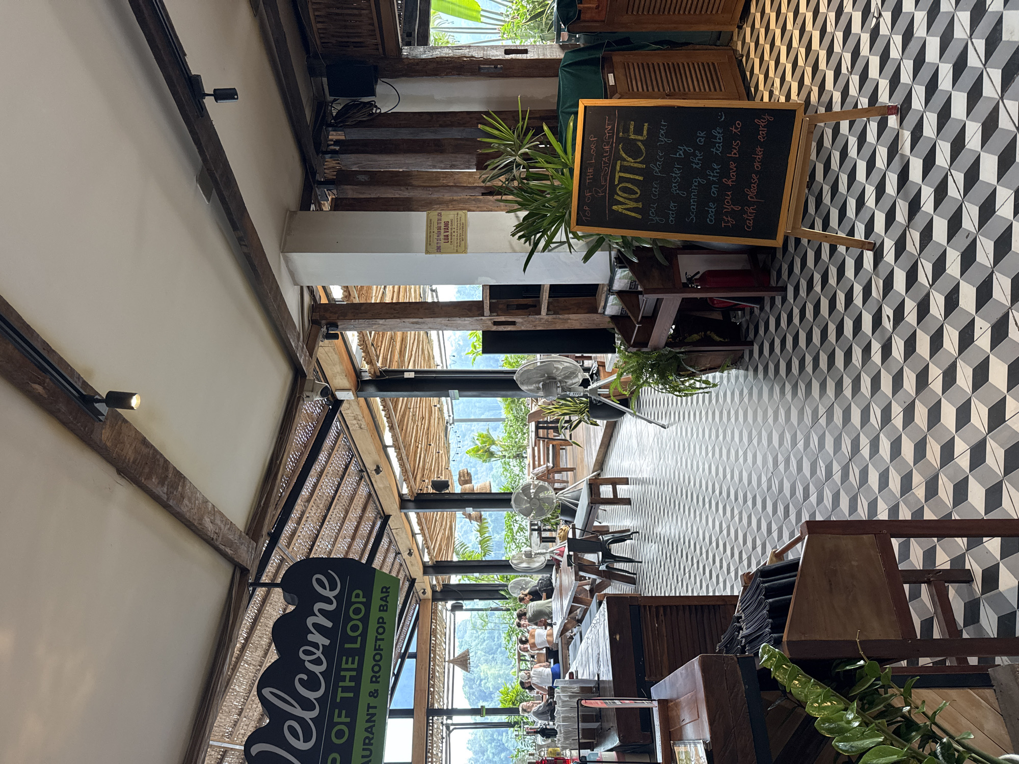 The image depicts the interior of "TOP OF THE LOOP RESTAURANT & ROOFTOP BAR" located in Hà Giang 1 Ward, Vietnam. The scene captures a spacious, open-concept dining area during the daytime, characterized by a rustic and natural design.
The setting features prominent wooden beams and columns supporting a textured, possibly woven bamboo or wicker, ceiling. Several white ceiling fans are suspended, suggesting a warm climate. The floor boasts a distinctive black, white, and grey geometric patterned tile that creates a three-dimensional cube effect. Lush green plants are interspersed throughout the venue, complementing the natural light streaming in from large windows or open sections along the far wall.
In the mid-ground and background, several people are seated at wooden tables and chairs, appearing to be dining or relaxing. Beyond them, a panoramic view of verdant mountains and dense tropical foliage is visible, emphasizing the scenic location.
Notable textual elements include a large green and white sign on the left that reads: "Welcome TOP OF THE LOOP RESTAURANT & ROOFTOP BAR." On the right, a black chalkboard easel displays a "NOTICE" in white chalk: "you can place your order faster by scanning the QR code on the table. If you have bus to catch please order early." Additionally, a smaller yellow sign mounted on a white wall section contains Vietnamese text: "CÔNG TY CỔ PHẦN ĐẦU TƯ DU LỊCH LÚA VÀNG CHI NHÁNH HÀ GIANG," which translates to "LUA VANG TOURISM INVESTMENT JOINT STOCK COMPANY HA GIANG BRANCH." The overall atmosphere is bright, open, and inviting, suggesting a popular spot for enjoying food and views.