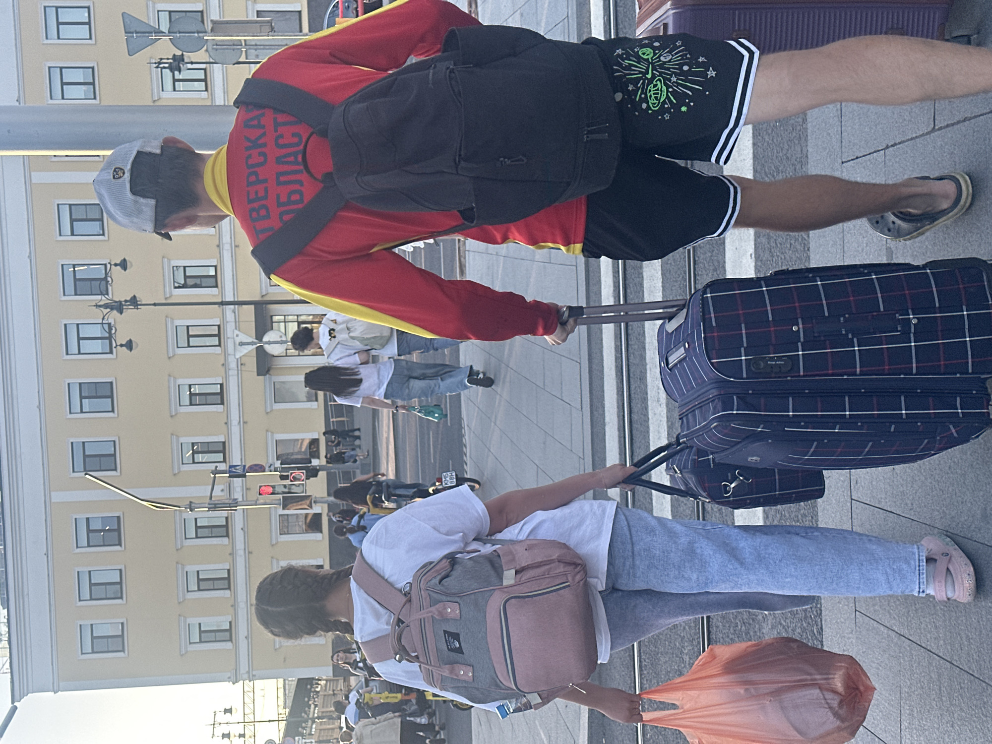 In this vertical, eye-level shot, a group of people are seen walking on a paved surface in front of a light-colored building. The sun appears to be low in the sky, casting soft shadows and creating a warm, late afternoon or early morning light.
In the foreground, a person wearing a red and yellow jacket with black trim and a black backpack walks away from the camera, pulling a suitcase. The jacket has text printed on the back in black lettering that appears to say "ТВЕРСКАЯ ОБЛАСТЬ" (Tver Oblast). They are also wearing black shorts with white stripes and black flip-flops.
To the right of this person, another individual is pulling a blue and red plaid suitcase. This person is wearing a white t-shirt and light blue jeans. They are also carrying a pink and gray backpack and an orange plastic bag.
In the background, more people are visible walking, some with luggage. The building behind them has multiple windows and architectural details. Traffic lights and streetlights are also present, indicating an urban environment. The overall scene suggests people are traveling or perhaps arriving at a destination.