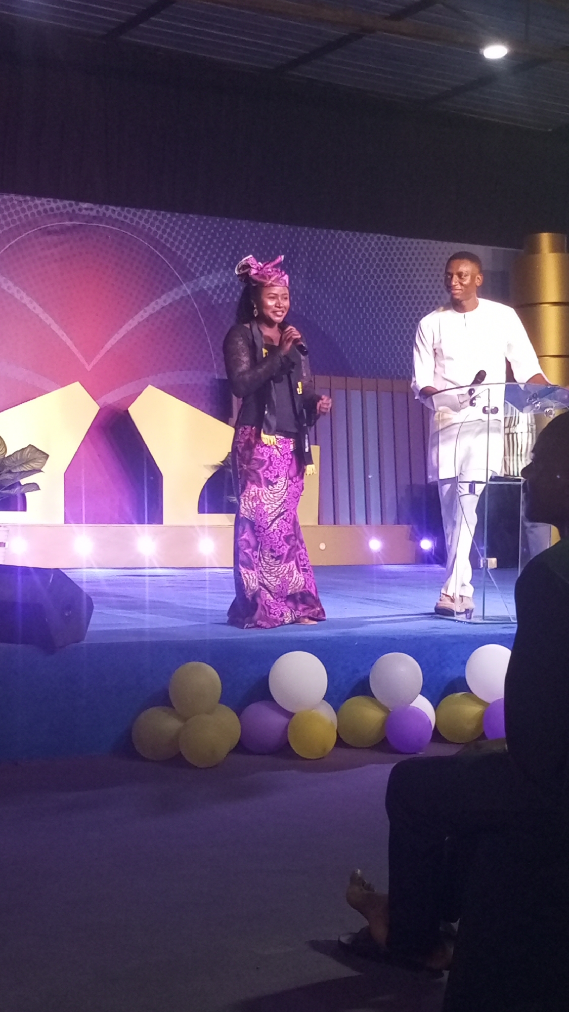 A woman wearing a pink headwrap and a patterned purple skirt stands center stage, holding a microphone and speaking. To her right, a man in a white outfit stands behind a clear podium, facing her. The stage is lit with colorful spotlights and decorated with a large, stylized heart graphic on the backdrop. Balloons in yellow, purple, and white are scattered on the floor in front of the stage. The event appears to be taking place indoors, possibly in a church or event hall, in Durumi, Nigeria.