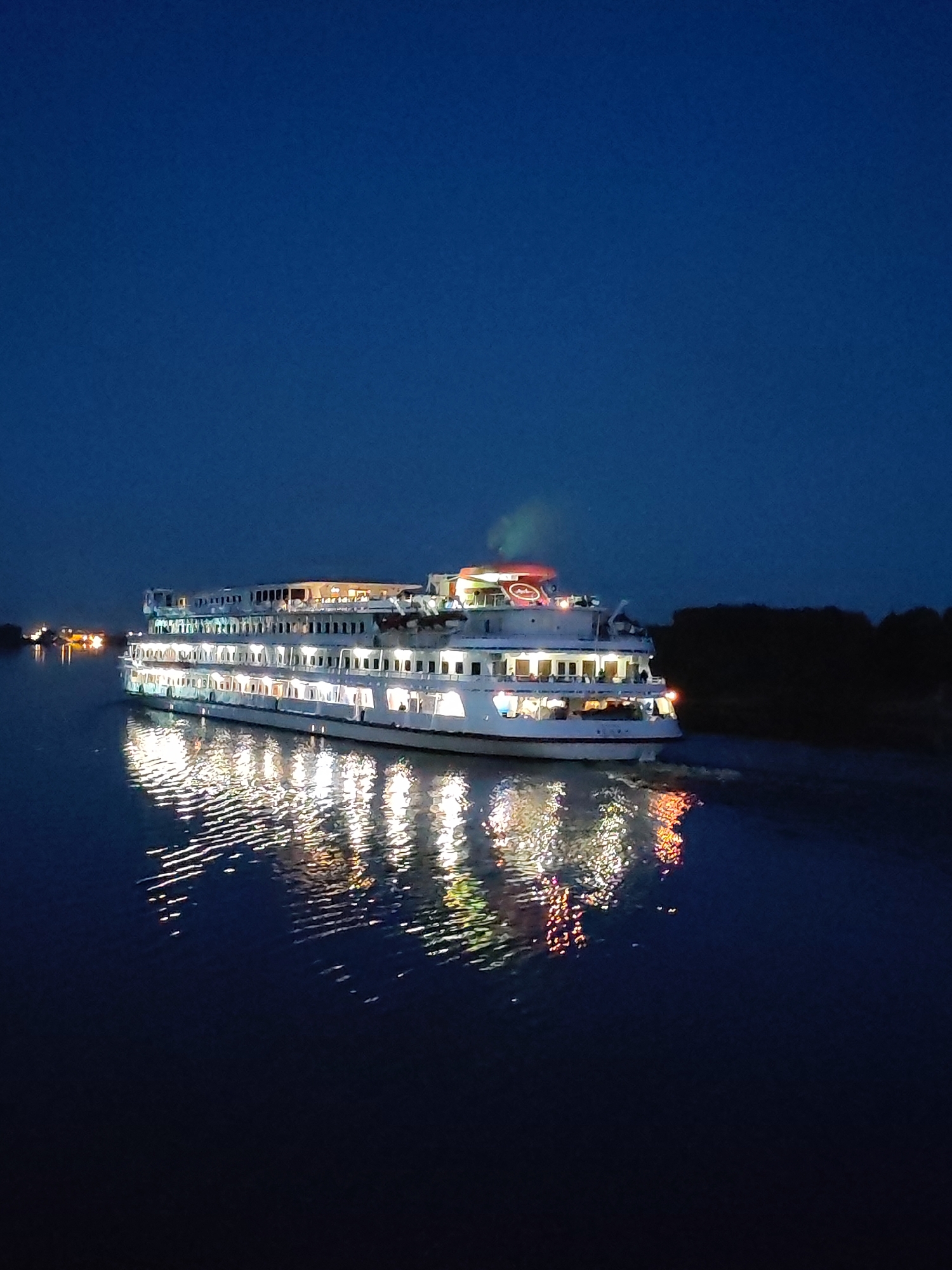 The image shows a large, multi-decked white riverboat illuminated by interior and exterior lights, navigating a dark body of water at night. The boat is positioned in the center of the frame and is moving from left to right. Its windows glow with warm light, and reflections of these lights shimmer on the water's surface. A plume of light green smoke or steam is visible emanating from the top deck, near the front of the vessel. In the background, on the right side of the image, a dark, tree-lined shore is visible. On the left side, distant lights suggest other vessels or structures. The sky is a deep, clear blue, indicating twilight or nighttime. The location is identified as the city of Bol'shie Mkhi, Russia.