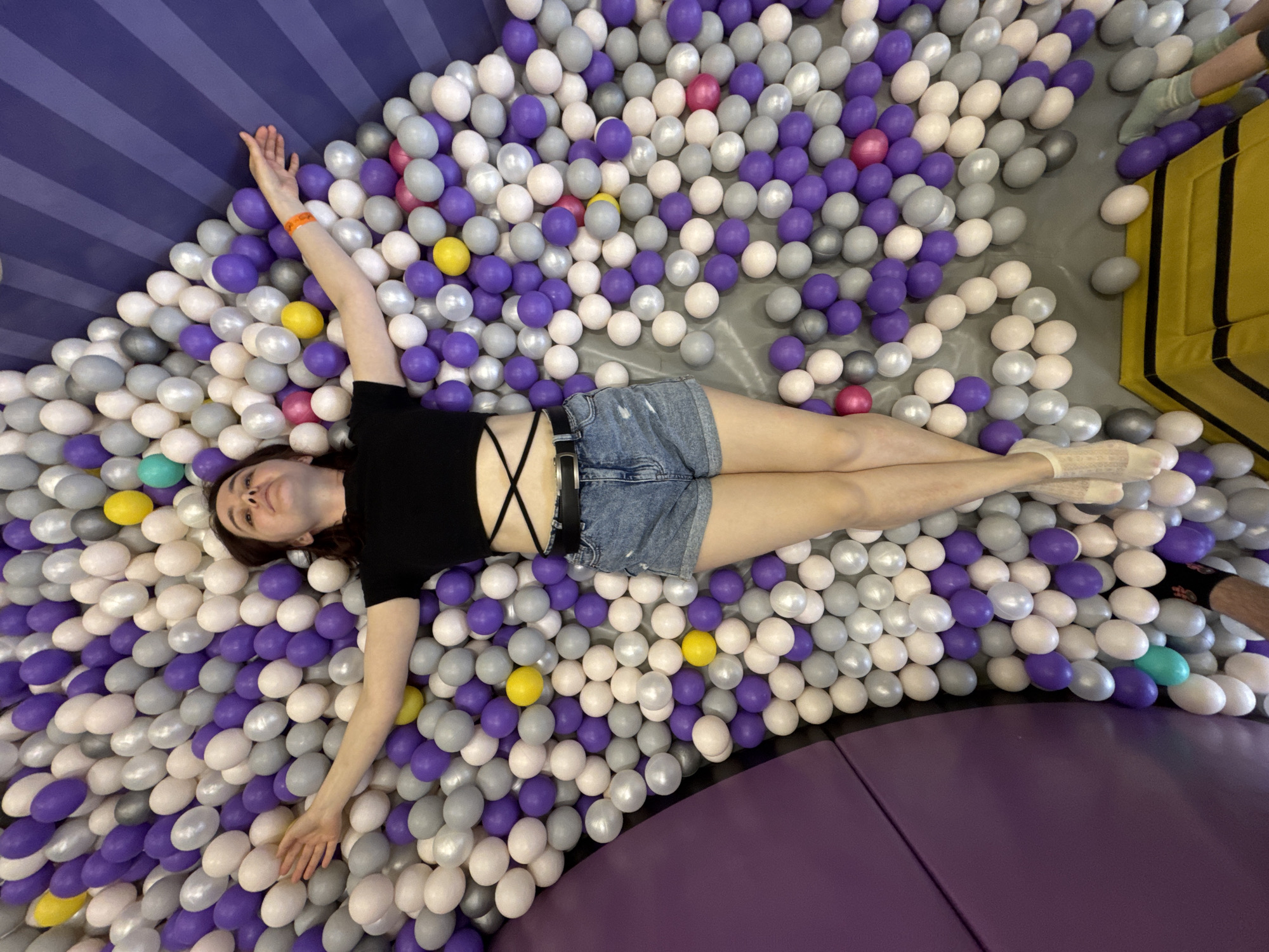 A young woman is lying in a ball pit filled with white, gray, and purple plastic balls, with accents of yellow, pink, and teal. She is wearing a black crop top with criss-cross straps and denim shorts. Her arms are outstretched. The ball pit is located within an indoor playground, with padded walls and flooring in shades of purple. The photograph may be taken in Sochi, Russia, given the request for the location in the prompt.