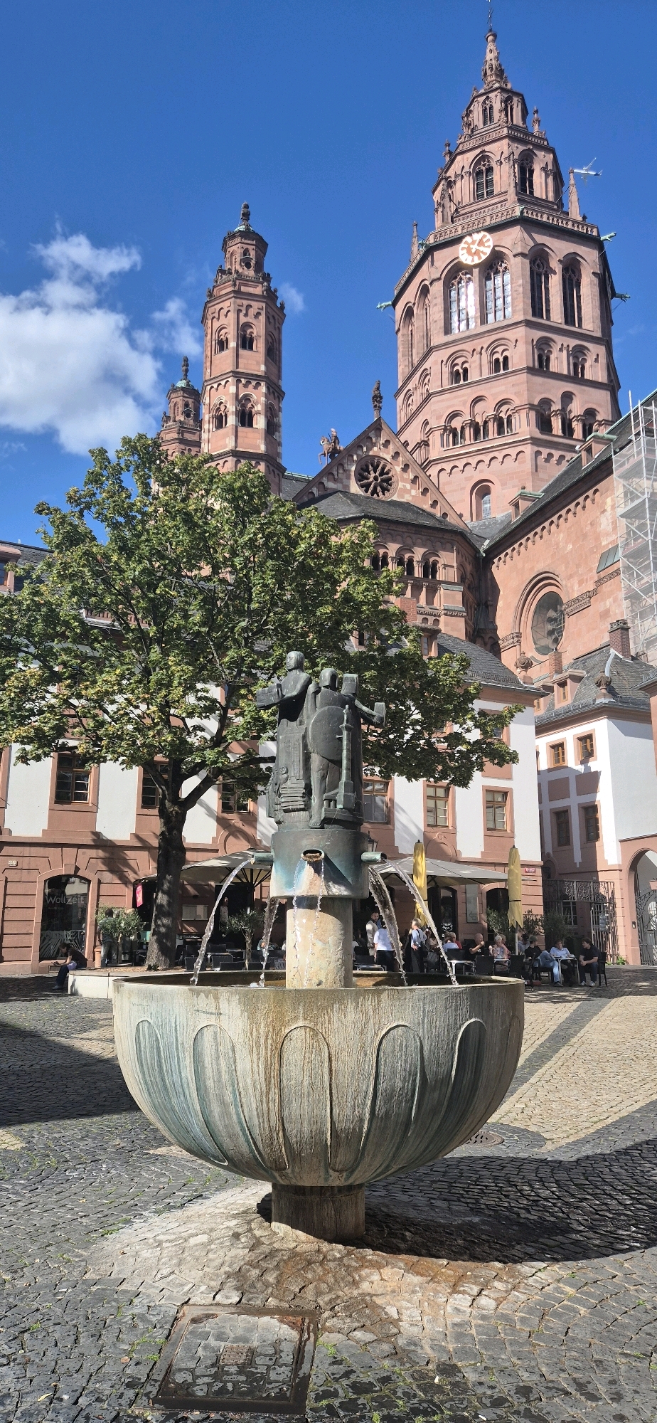 The image depicts a sunny day in a cobblestone square in front of the Mainz Cathedral (Mainzer Dom), located in Mainz, Germany. In the foreground, a large, aged stone fountain with a bronze sculpture of three figures at its center is active, with water cascading from its spouts. The figures in the sculpture appear to be holding shields and staffs. Behind the fountain, a large, leafy green tree stands, partially obscuring the ornate red sandstone architecture of the cathedral, which features multiple towers and arched windows. A few scattered people are visible sitting at tables outside a cafe or restaurant to the right of the tree and in the middle ground. The sky above is a clear blue with a few wispy white clouds. The overall scene is one of historical architecture and a lively public space on a bright day.