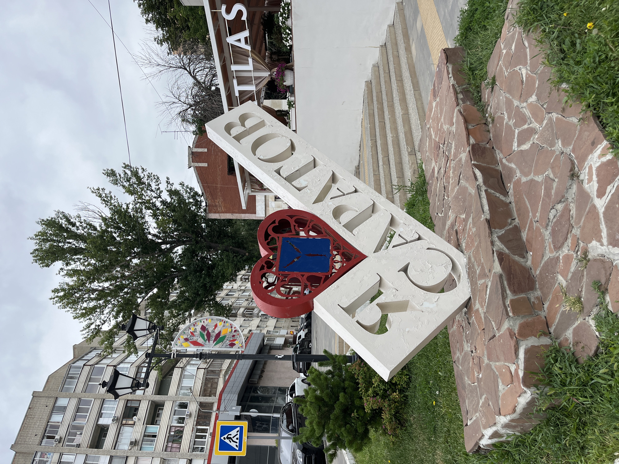 The image shows a monument in Saratov, Russia, that spells out "Я С АРАТОВ" (Ya Saratov - I am Saratov). The monument is made of large, white, three-dimensional letters, angled upwards and to the right. The first letter, "Я", is on the left, followed by "С" and then "АРАТОВ". In the center of the "С" is a red heart cutout containing a blue emblem with a square and three triangular shapes. The monument stands on a tiered base made of irregularly shaped reddish-brown stones. To the left of the monument is a green bush, and further left are a street lamp with two black lamps and a decorative stained-glass panel, a pedestrian crossing sign, and several parked cars. In the background, a multi-story apartment building with many windows is visible, along with trees and a red brick building with a white awning that reads "ATLAS" in white letters. The sky is overcast.