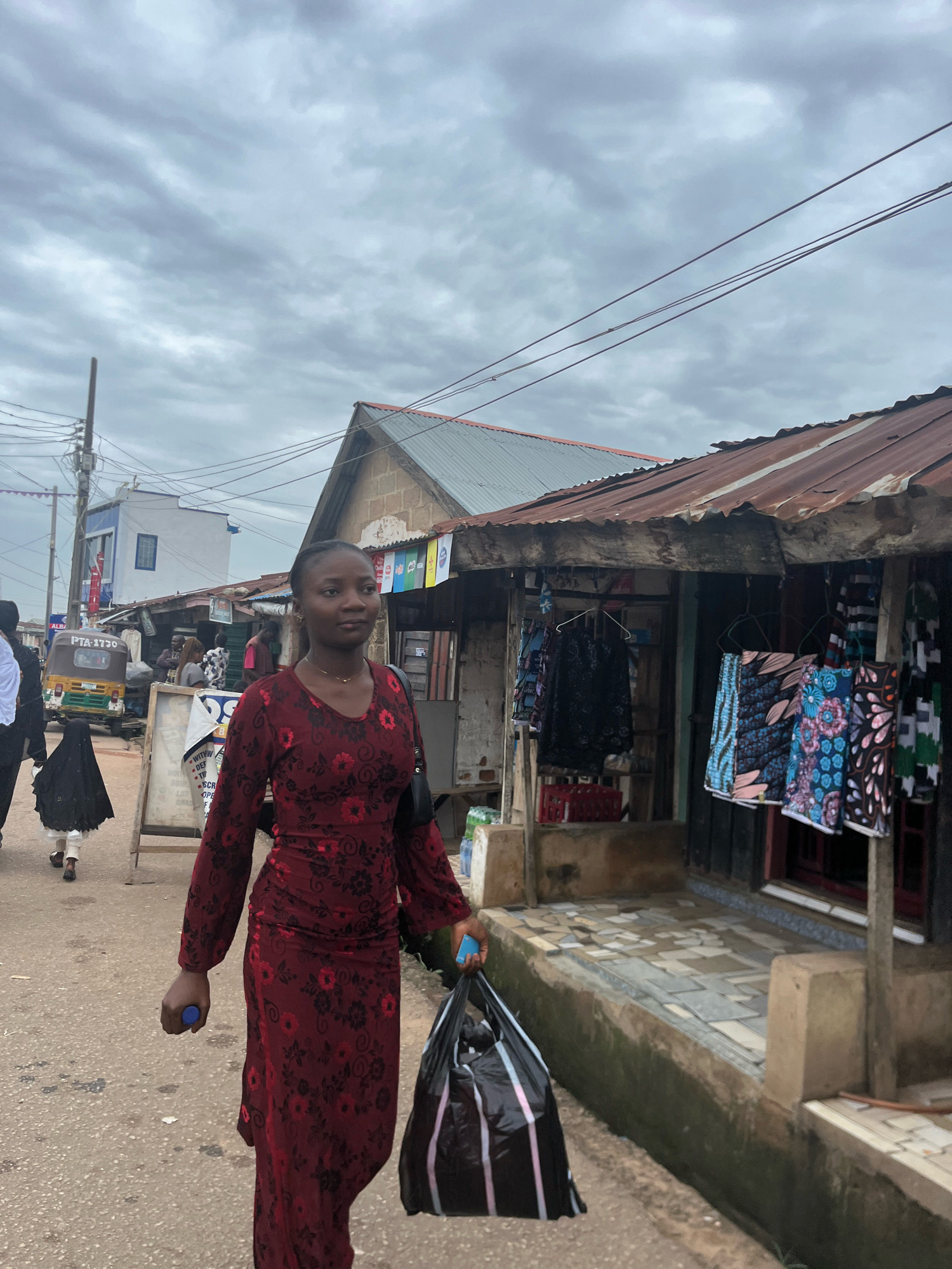 The image captures a street scene in Jos, Nigeria, under a cloudy sky. A young woman in a long, patterned red dress walks towards the viewer, carrying a black plastic bag in her right hand and holding a blue object in her left hand. She is on a paved street, with a row of market stalls on her right. The stalls have corrugated metal roofs and display colorful fabrics and clothing. In the background, a yellow and green three-wheeled vehicle, a "Keke," is visible, along with other pedestrians and buildings. The overall impression is of a busy marketplace in an urban environment.
