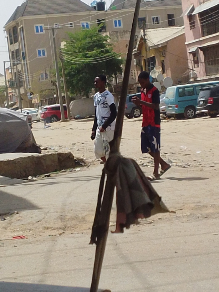 The image depicts a daytime street scene in Sabon Gari East, Nigeria. In the immediate foreground, a dark vertical pole with a collapsed, dark, tattered fabric object (possibly an umbrella) attached to it occupies the lower center. Two males are walking from left to right across the midground. The individual on the left wears a white t-shirt with dark patterns, black long sleeves, and light-colored shorts, looking straight ahead. The individual on the right wears a red and black short-sleeved shirt with a white logo, blue and white patterned shorts, and white slide sandals; he is observed looking down at a mobile phone held in both hands. The ground consists of uneven dirt and paved sections. In the background, a light-colored multi-story building with blue-framed windows and a water tank on its roof is visible on the left, while other buildings, including one with satellite dishes, line the right. Several vehicles, including a red SUV, a light blue van, and a dark SUV, are visible parked or in motion further down the street. The scene is brightly lit by direct sunlight.
