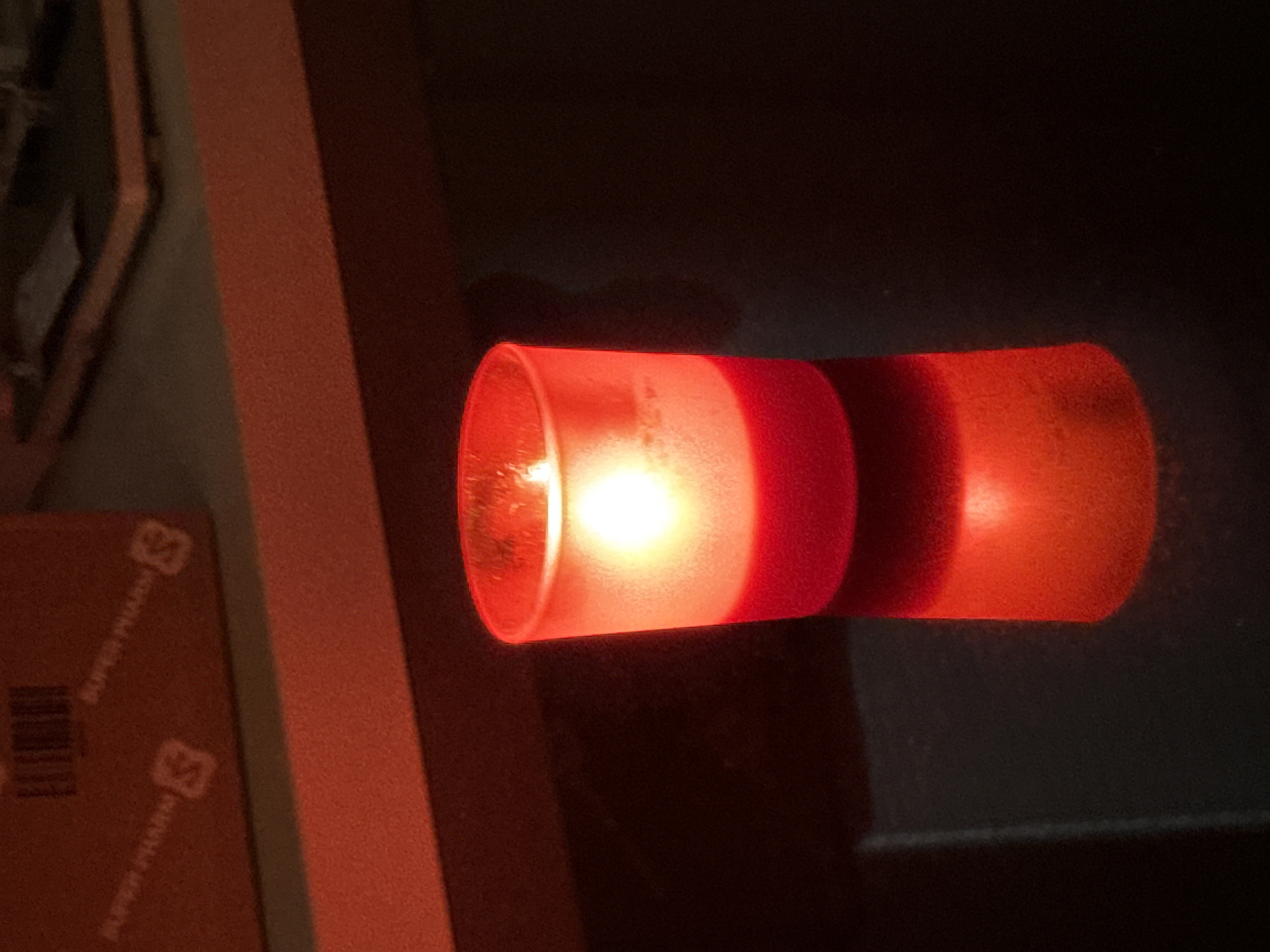 A red votive candle flickers in a glass holder. The candle is lit and casts a warm glow. The holder is cylindrical and made of frosted glass, with a red base. The candle sits on a dark, reflective surface, possibly a table. In the background, a red surface with some printed text is visible, along with a blurred out structure. The overall scene is dimly lit, with the candle being the primary light source. The location is Unknown, Unknown.