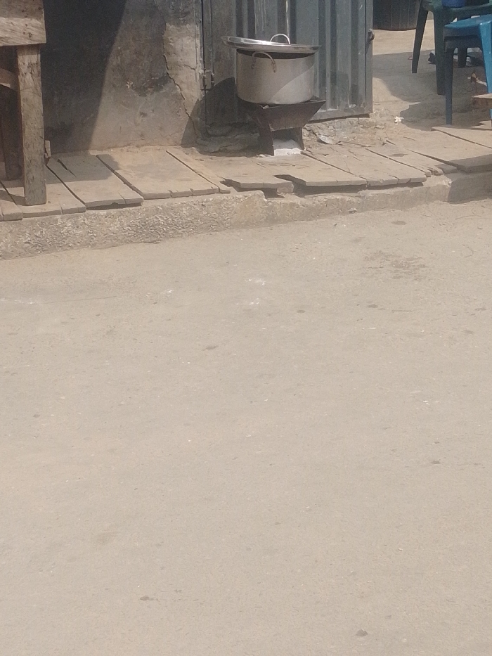 The scene is a ground-level view of an outdoor urban environment in Agani, Nigeria, during daylight. The foreground consists of a light-colored, possibly unpaved dirt or gravel road surface with minor irregularities and debris. In the midground, a raised pedestrian path constructed from multiple light-colored wooden planks runs horizontally across the frame, forming a transition from the road to a building structure.
On this wooden plank path, a large, silver-colored aluminum pot with a matching lid rests on a dark-colored, rudimentary metal cooking stand or charcoal stove. This stove is positioned directly in front of a weathered, light-colored wall exhibiting visible cracks and damage. Immediately to the right of the wall section is a dark, corrugated metal door or gate, which appears slightly ajar. To the far left, a vertical wooden beam, likely part of a table or stall structure, is partially visible. Further to the right in the background, a portion of a blue plastic chair and a dark, indistinct object are present. No individuals are depicted in the frame.