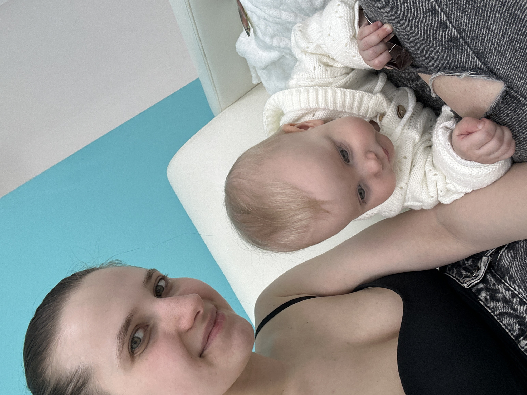 An indoor scene depicts an adult female and an infant. The woman is positioned on the left, looking directly at the camera with a slight smile. She has dark hair pulled back and wears a black strappy top and dark grey ripped jeans with a visible tear on the knee. The infant, with light blonde hair, is seated or crawling on the woman's lap or alongside her. The infant wears a white knitted cardigan with light brown buttons and is looking downwards and slightly to the left, holding a dark, rectangular object in its right hand. A white upholstered sofa is visible behind the subjects. The background wall features a diagonal color division, with the upper left section colored teal and the upper right section white. A white knitted blanket or fabric is visible on the sofa to the right of the infant. The specific location within the city of Moscow, Russia, is not identifiable from the visual evidence.