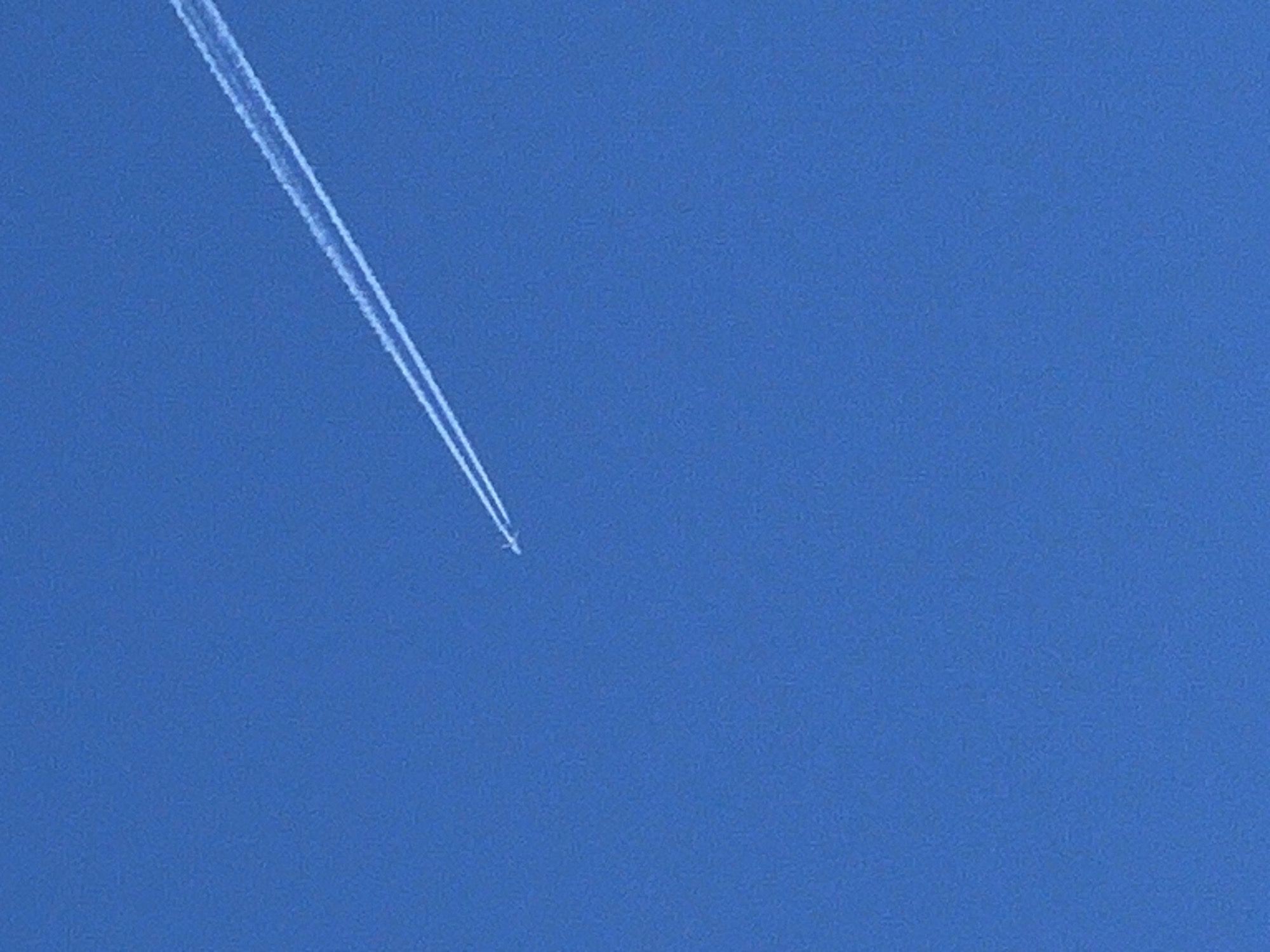 A clear, blue sky fills the frame. An airplane flies diagonally from the upper left to the center, leaving a white contrail behind it. The airplane is small and distant. There are no people, buildings, or other objects visible other than the airplane and its contrail against the sky. The location and the city are not visibly or directly identifiable in this image.