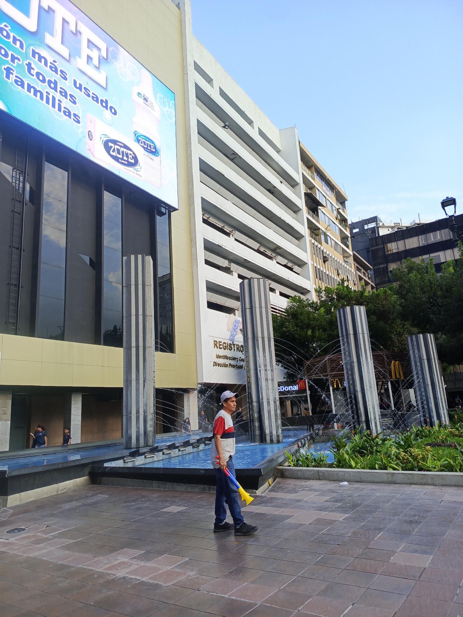 The image depicts a street scene in Guayaquil, Ecuador, likely in an urban commercial area. In the foreground, a man wearing a white, red, and blue striped shirt, blue jeans, and a white baseball cap walks on a paved area, holding a yellow and blue toy horn. He is walking towards the right side of the frame.
In the mid-ground, there is a water feature with several tall, fluted pillars through which water sprays. Behind the water feature, a building with large windows displays a prominent advertisement for "ZOTE" soap and detergent on a large digital screen. The advertisement features images of Zote products and text in Spanish, including "rón más usado por todas familias" (most used by all families). Adjacent to the Zote advertisement, another building is visible, with multiple floors and windows. Further back, there are signs for McDonald's.
In the background, several other buildings of varying heights and architectural styles are visible against a clear blue sky. Some of the buildings appear to be modern office buildings or residential complexes. The overall atmosphere suggests a daytime scene in a bustling city.