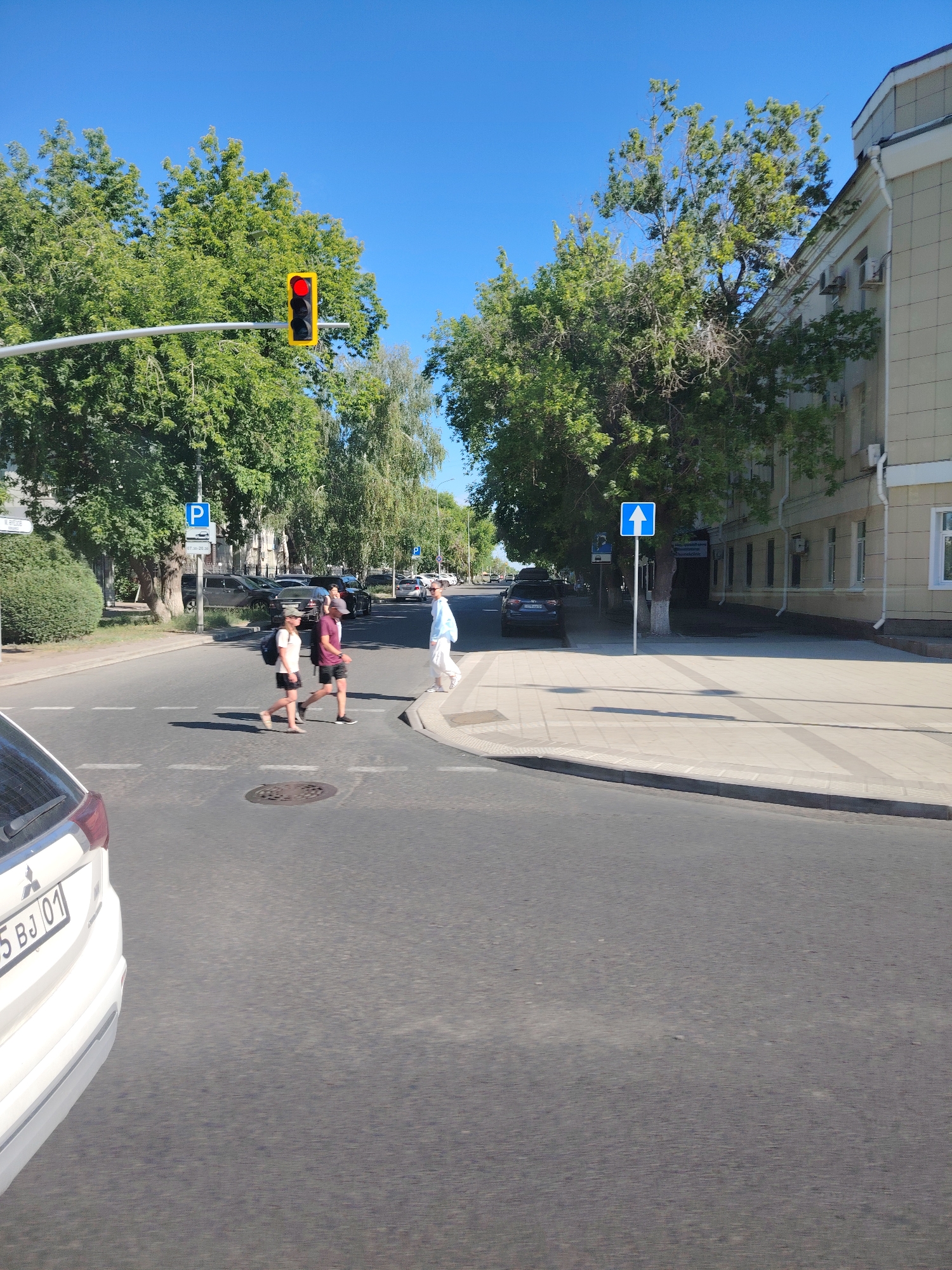 The image depicts a street scene in Astana, Kazakhstan, under a clear blue sky. In the foreground, the rear of a white car is visible on the right. The street features a pedestrian crossing. Three individuals are crossing the street from left to right: two younger people, one wearing a white t-shirt and a backpack, and another in a dark t-shirt and shorts, both with hats; and a third person, further ahead and to the right, wearing a light blue outfit and carrying a white bag. A traffic light is visible on a pole, showing a red light. Traffic signs are also present, including a blue sign with a white "P" indicating parking, and a blue sign with a white upward-pointing arrow. Trees line the street, and a modern building with a light-colored facade is visible on the right side of the frame. The overall environment suggests a sunny day in an urban setting.