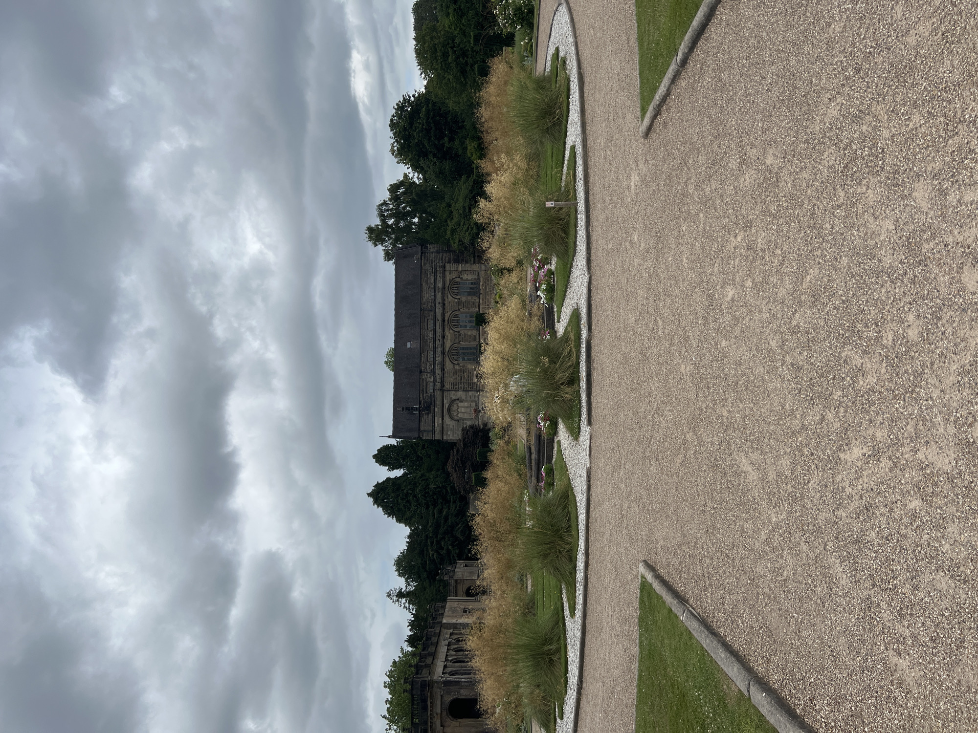 The image shows a view of a historic building, likely a church or cathedral, with intricate stone architecture. The building is surrounded by lush green grass, mature trees, and decorative ornamental grasses. A pathway made of gravel is prominent in the foreground, curving to the right and bordered by grass and stone. The sky is overcast with dramatic, heavy grey clouds, suggesting a cool or temperate climate.
The scene appears to be a well-maintained garden or park area adjacent to the historic structure. There are no people visible in the image, and the activity taking place is simply the presentation of the landscape and architecture. Notable details include the textured stone of the building, the feathery appearance of the ornamental grasses, and the light-colored gravel path. The overall mood is serene and somewhat somber due to the cloudy sky.
The location context is Stafford, United Kingdom, which aligns with the architectural style and the greenery. There is no visible text in the image. The time of day is difficult to ascertain precisely due to the overcast conditions, but it appears to be daytime.