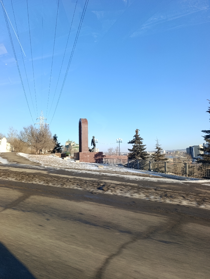 The image displays the Andrey Dubensky Monument in Krasnoyarsk, Russia, situated on an elevated terrain near Peace Square and the Krasnoyarsk Regional Museum of Local Lore. The monument features a tall, reddish-brown rectangular stone column with a dark-colored statue of a man, Andrey Dubensky, positioned to its right. The statue depicts a standing figure, appearing to hold an object in his left hand while gesturing with his right.
Behind the monument and to the left, a multi-story building with a distinct green roof and spire, identified as the Krasnoyarsk Regional Museum of Local Lore, is partially visible amidst bare deciduous trees. To the monument's right, several evergreen conifer trees and a dark metal fence delineate the edge of the elevated area. In the foreground, a dark, partially snow-covered road surface, exhibiting signs of motion blur, occupies the lower portion of the frame. Multiple power lines traverse the clear blue sky diagonally from the upper left towards a metal lattice power pylon situated mid-ground left of the monument. Distant urban structures are visible on the horizon. The scene is illuminated by direct sunlight, indicating a cold season with patches of snow on the ground. No individuals are visibly present in the scene aside from the statue.