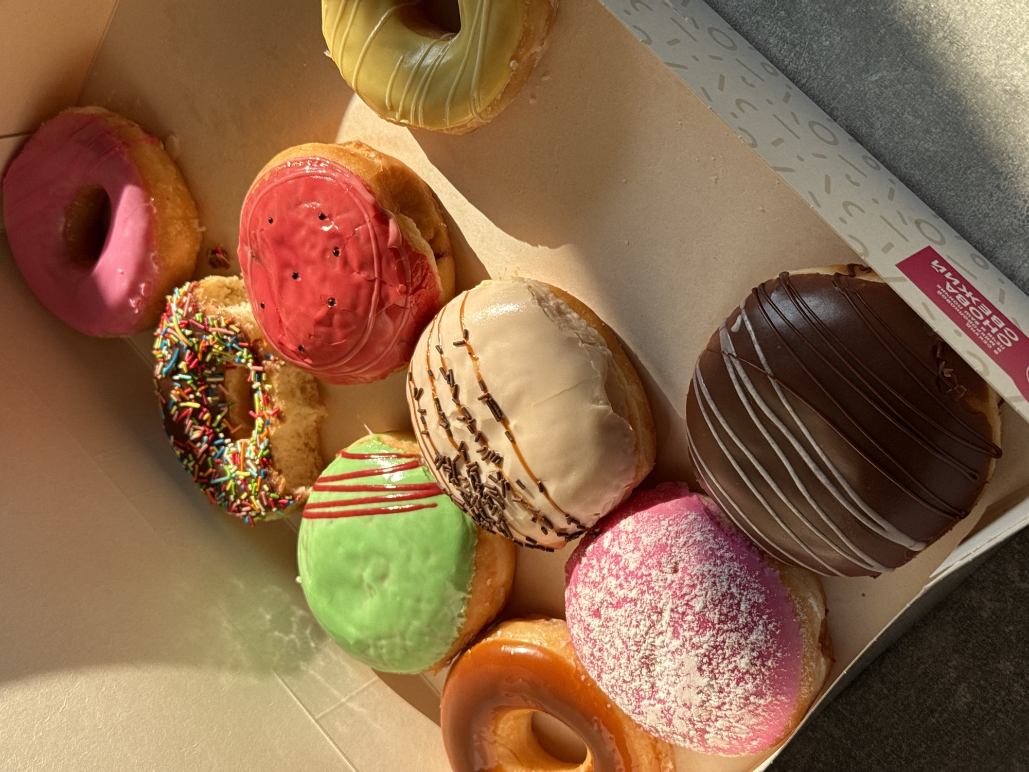 A box of assorted donuts is displayed. The donuts have various toppings including pink frosting with powdered sugar, caramel glaze, green frosting with red stripes, a red "watermelon" style frosting, chocolate frosting with white stripes, and sprinkles. The box is angled and casts shadows from the sunlight hitting it from the left. The background includes a textured wall and a grey surface. The location is a box of donuts in Lyubertsy, Russia, though no people are present in the image.