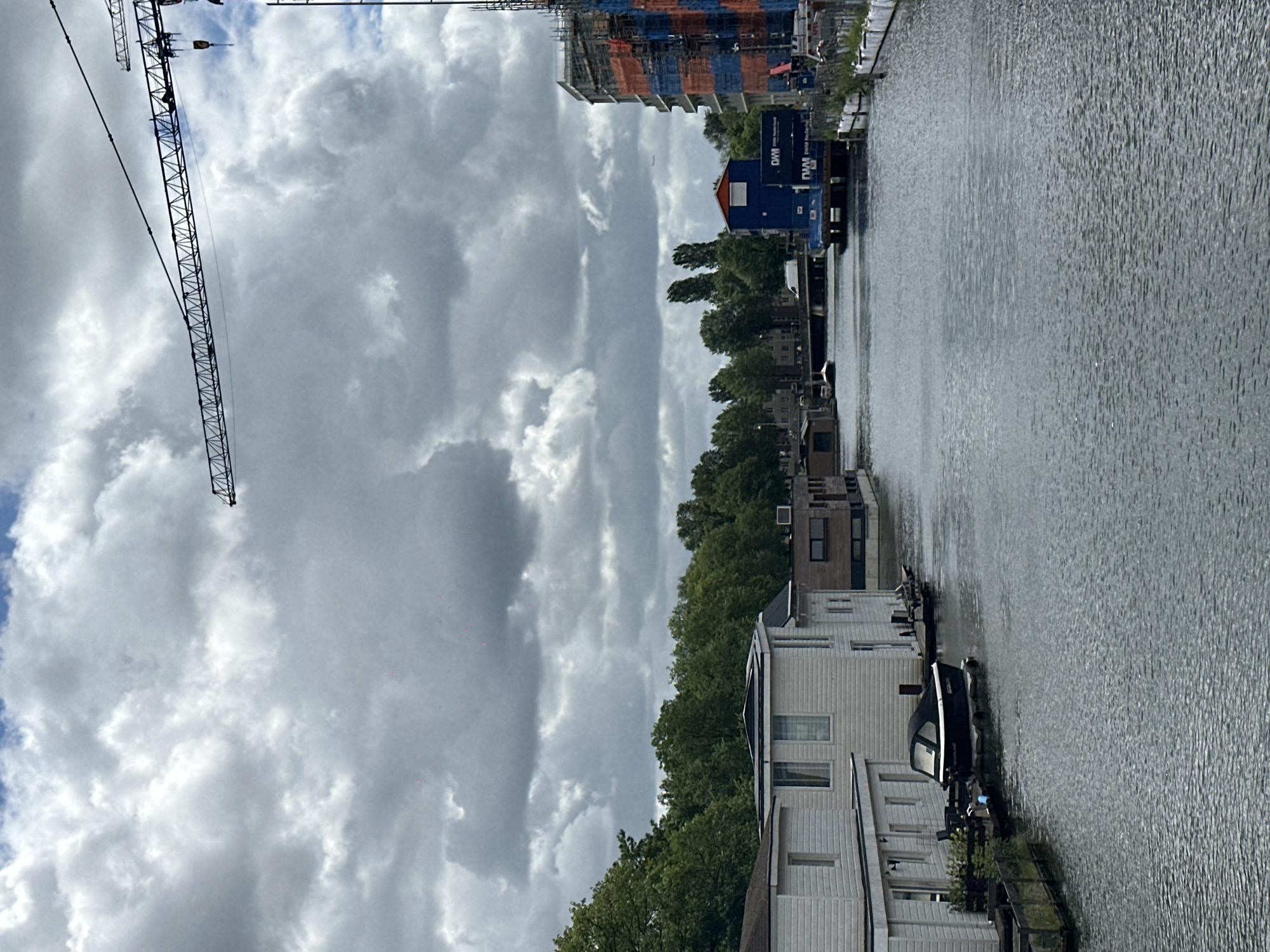 The image depicts a canal scene in Amsterdam, Netherlands, with a cloudy sky. In the foreground, the water of the canal is calm. On the left bank, several white houseboats are moored, one with a black boat docked next to it. Further down the canal, on the right bank, a blue structure with "DWM" branding is visible, adjacent to a multi-story building under construction, with scaffolding and orange and blue sheeting. Trees line the background on the left bank, and a bridge crosses the canal in the distance. A construction crane is visible in the upper right quadrant of the image, against the cloudy sky.
