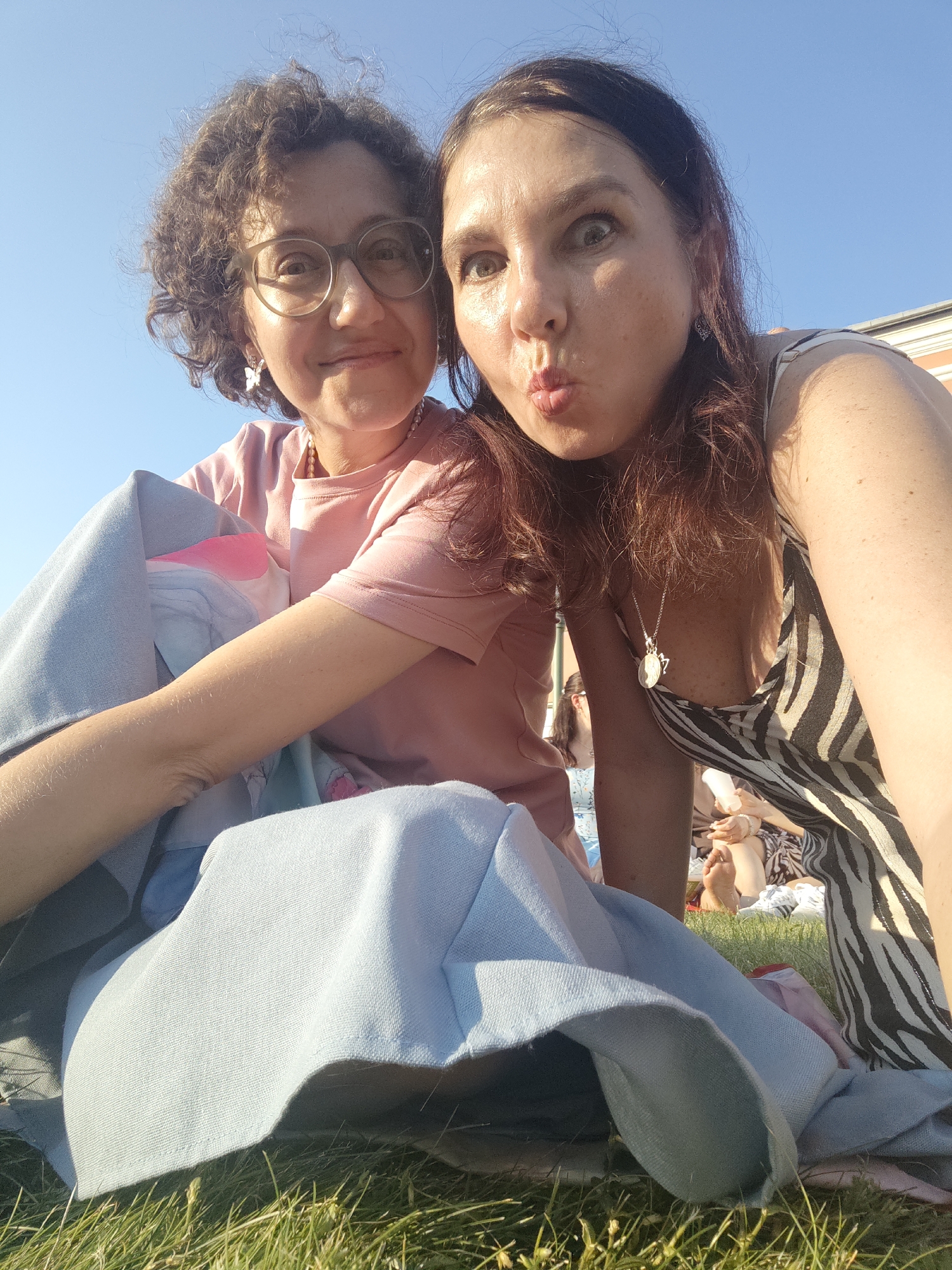 This is a selfie taken outdoors on a sunny day. Two women are the main subjects, positioned in the foreground and looking at the camera. The woman on the left has curly brown hair, wears glasses and a pink t-shirt. She is looking directly at the camera and smiling. The woman on the right has long brown hair and is wearing a black and white striped dress. She is making a "fish face" expression. Both women are sitting on grass, with a blue blanket draped over them and the woman on the left's lap. In the background, other people can be seen sitting on the grass, suggesting a gathering or event. The sky is clear and blue. The lighting suggests it is late afternoon or early evening. While the specific location within Saint Petersburg is not identifiable from the image alone, the presence of an outdoor grassy area with other people suggests a public park or a similar recreational space.