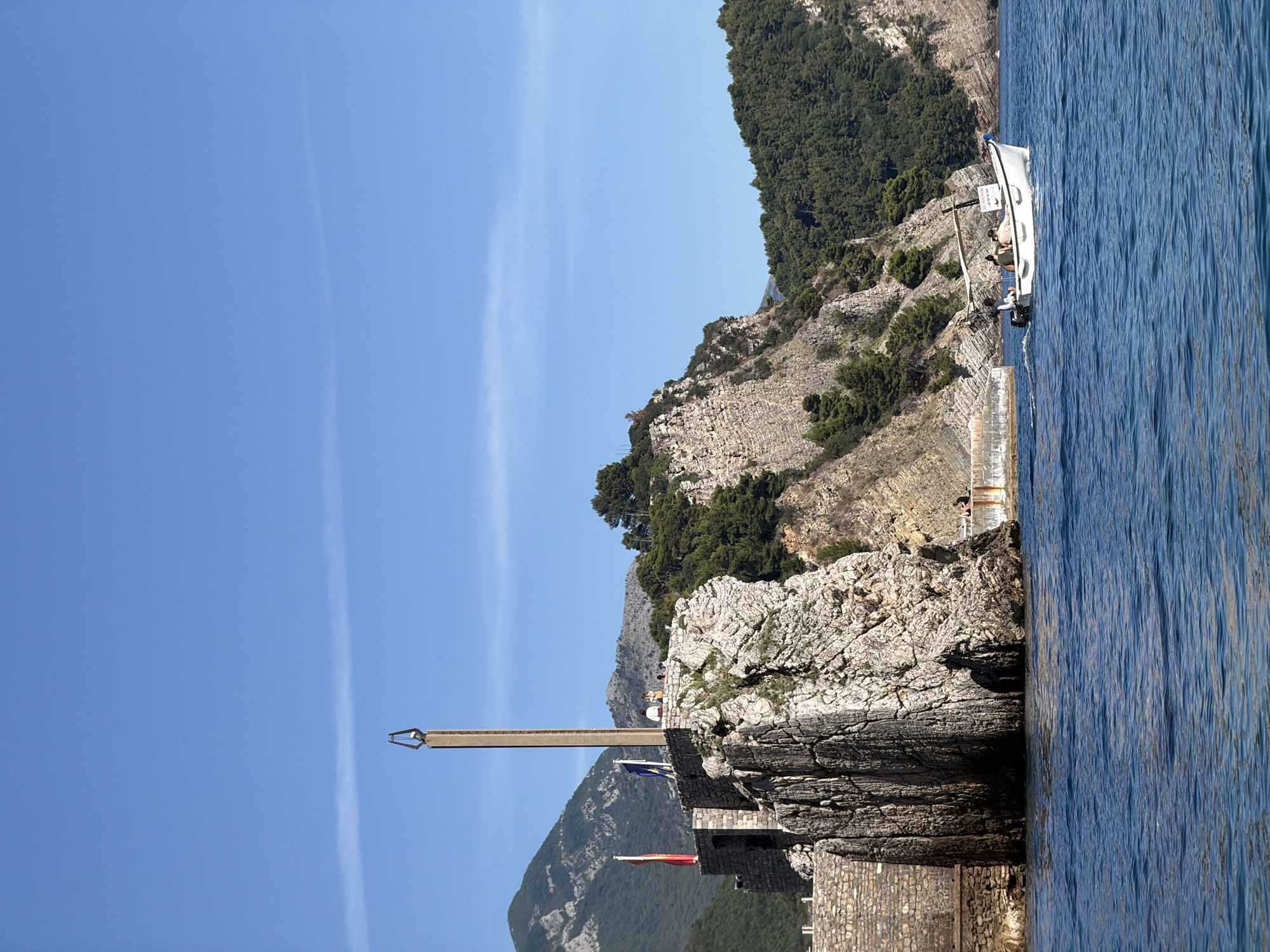 In Petrovac centar, Montenegro, a bright, clear blue sky is visible above a rugged coastal landscape. In the foreground, the deep blue sea ripples gently. A small white motorboat with a canopy is navigating the water, carrying several passengers. The boat has a visible registration or nameplate. To the left of the boat, a rocky cliff face rises steeply, topped by a stone structure with two flags – one red and one blue. Dominating the top of this structure is a tall, slender obelisk-like monument. Beyond the cliff, lush green trees cover the slopes of distant mountains, also under the clear blue sky. The overall scene depicts a serene coastal environment under sunny conditions.
