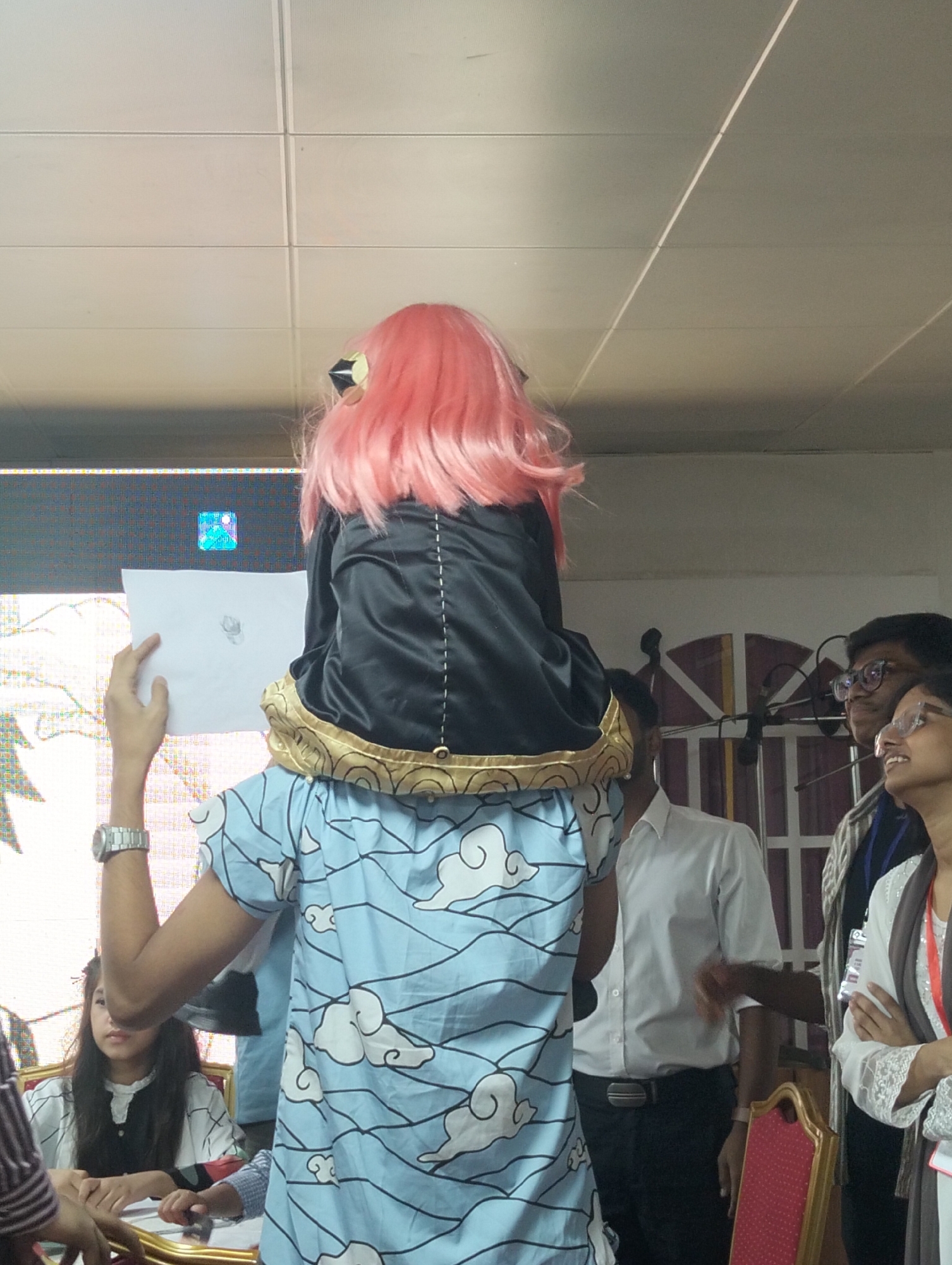 A person with a pink wig, possibly in costume, is being carried on the shoulders of another person. The person on the shoulders is holding a piece of paper with a drawing. The person carrying is wearing a light blue shirt with a cloud pattern. In the background, there are other people, some wearing glasses, and a large screen displaying an image. The environment appears to be indoors, possibly a convention or event, with a tiled ceiling.