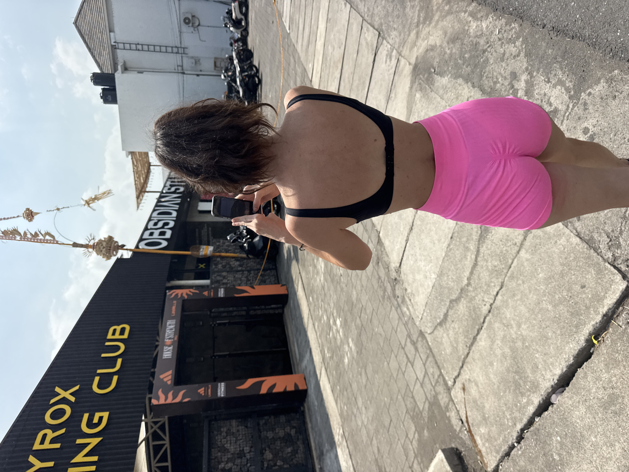 In Mengwi, Indonesia, a woman stands with her back to the camera, holding a smartphone to take a picture. She is wearing a black sports bra and bright pink shorts. Behind her, a building with the words "RYROX" and "ING CLUB" is visible, along with an entrance featuring orange and black banners. To the right of the building, the word "OBSIDIAN ST" is displayed. The ground is paved with rectangular stones, and a cracked concrete surface is in the foreground. In the background, several motorcycles are parked, and parts of other buildings and electrical infrastructure are visible under a clear sky.