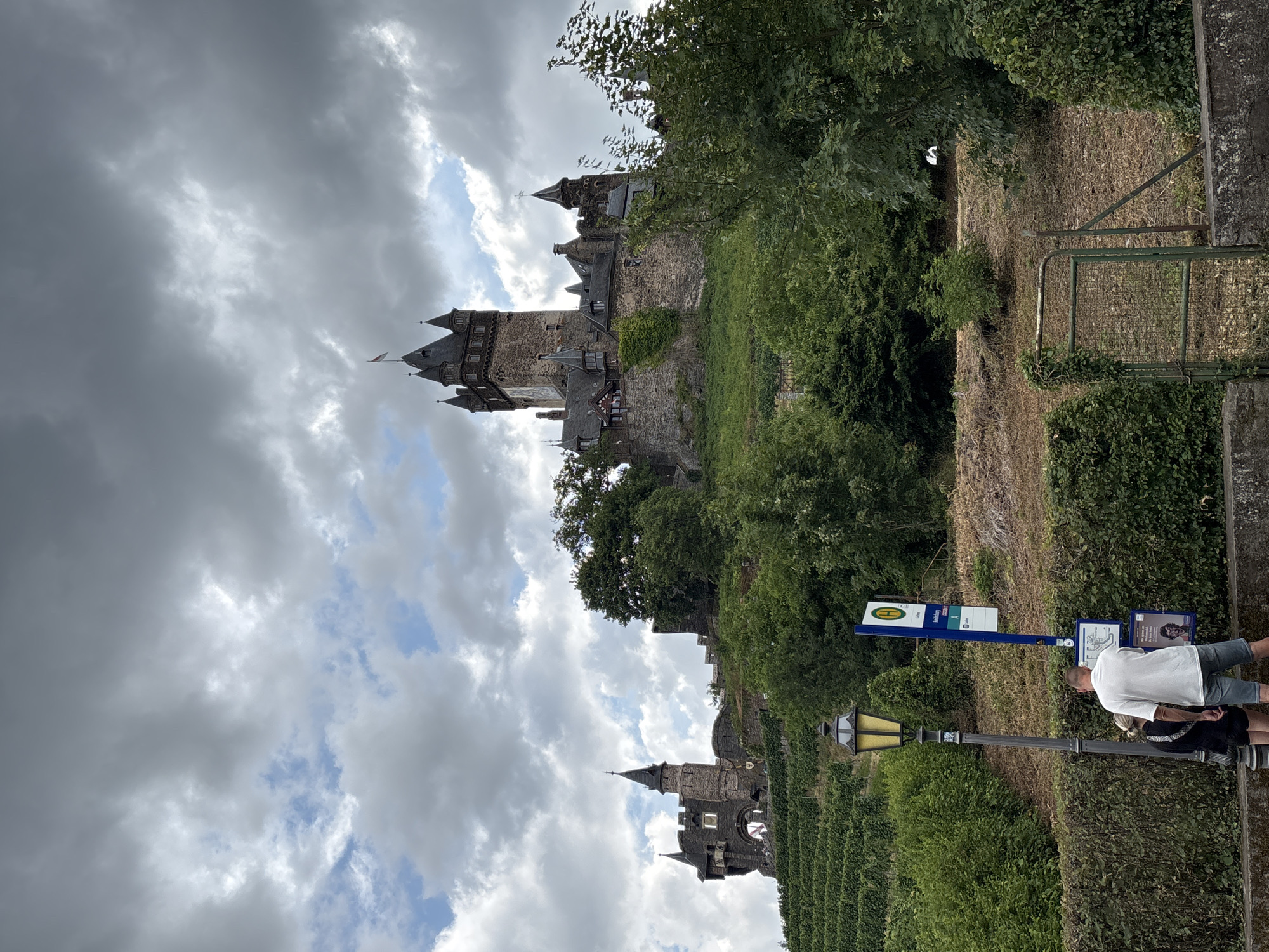 The image shows Reichsburg Castle, a prominent landmark in Cochem, Germany, perched on a hill overlooking the Moselle River. The castle is a large, medieval structure with multiple towers and turrets, and it is surrounded by lush green vineyards. In the foreground, a man and a woman are standing at a bus stop, waiting for transportation. The sky is overcast with dramatic clouds, suggesting a change in weather. A street lamp and a bus stop sign are also visible in the foreground. The overall scene conveys a sense of historical grandeur and rural charm.