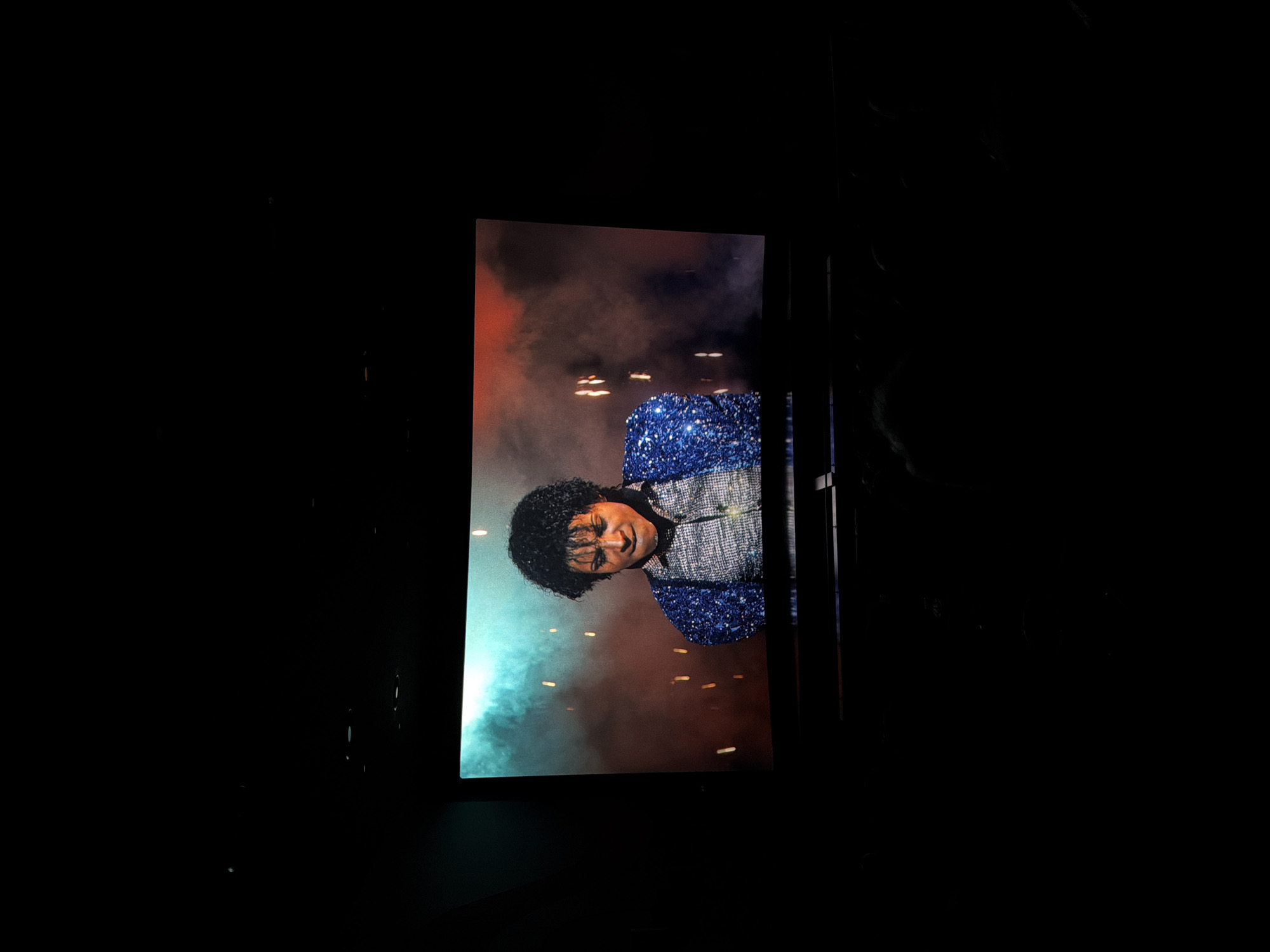 A large, vertically oriented screen dominates the central-right portion of the image, displaying an image of Michael Jackson. The rest of the scene is dark and indistinct, suggesting an indoor, low-light environment like a concert hall or a darkened room where a projection is being viewed.
Michael Jackson is shown from the chest up, his head slightly tilted. He has dark, curly hair and is wearing a shimmering jacket, which appears to be blue and silver, covered in sequins or glitter. His facial expression is a subtle smile or smirk. The background on the screen behind him is a vibrant mix of smoky red, orange, and blue-green hues, suggesting stage lighting or atmospheric effects. Scattered across this background are numerous small, bright, rectangular or oval lights, possibly reflections, stage elements, or digital effects, adding to a dynamic visual.
The image captures the moment of viewing this visual presentation of Michael Jackson, with the focus solely on the illuminated screen against the dark surroundings. No text is visible in the image.