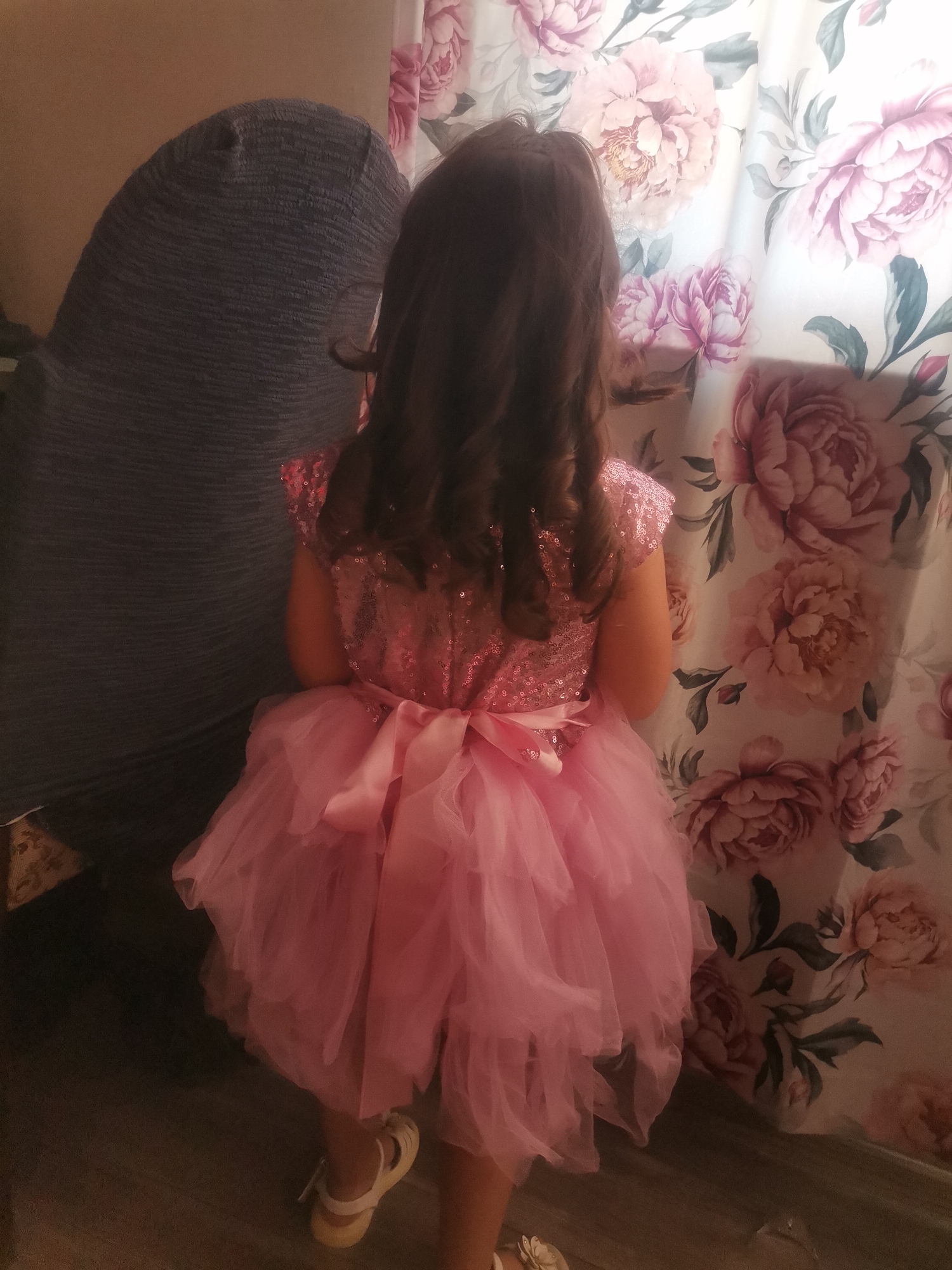 A full shot depicts a young girl, facing away from the viewer, dressed in a vibrant pink dress with a sparkly bodice and a full, layered tulle skirt. A ribbon bow is tied at the back of the dress. She is wearing white sandals.
To the left of the girl is a large, gray, textured object resembling a chair or a cushion. To her right is a curtain or backdrop adorned with a floral pattern of large, light pink flowers.
The flooring appears to be made of wood. The location is Unknown, Unknown.