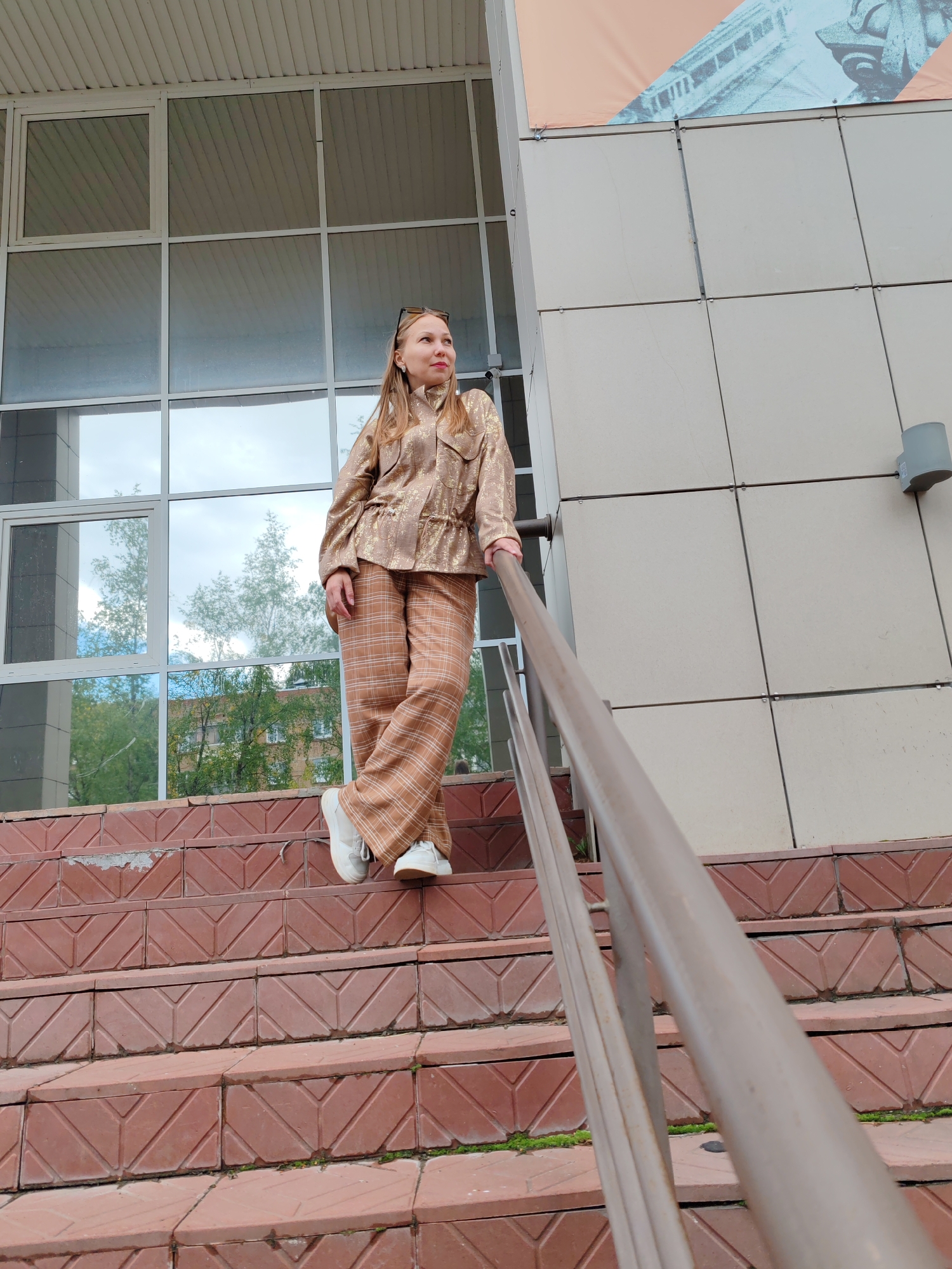 An adult female stands on a set of red brick stairs outside a modern building. The building has a large glass facade with reflections of trees and the sky. The woman is wearing a metallic gold jacket with a patterned drawstring waist and matching wide-leg plaid pants in shades of brown and white. She has white sneakers and sunglasses perched on her head. Her left hand rests on a metal railing that runs alongside the stairs. The stairs are made of textured red bricks arranged in a diamond pattern. There are small patches of moss growing between some of the bricks. The lighting suggests it is daytime. The architectural style of the building and the surrounding greenery are consistent with an urban environment in Syktyvkar, Russia.