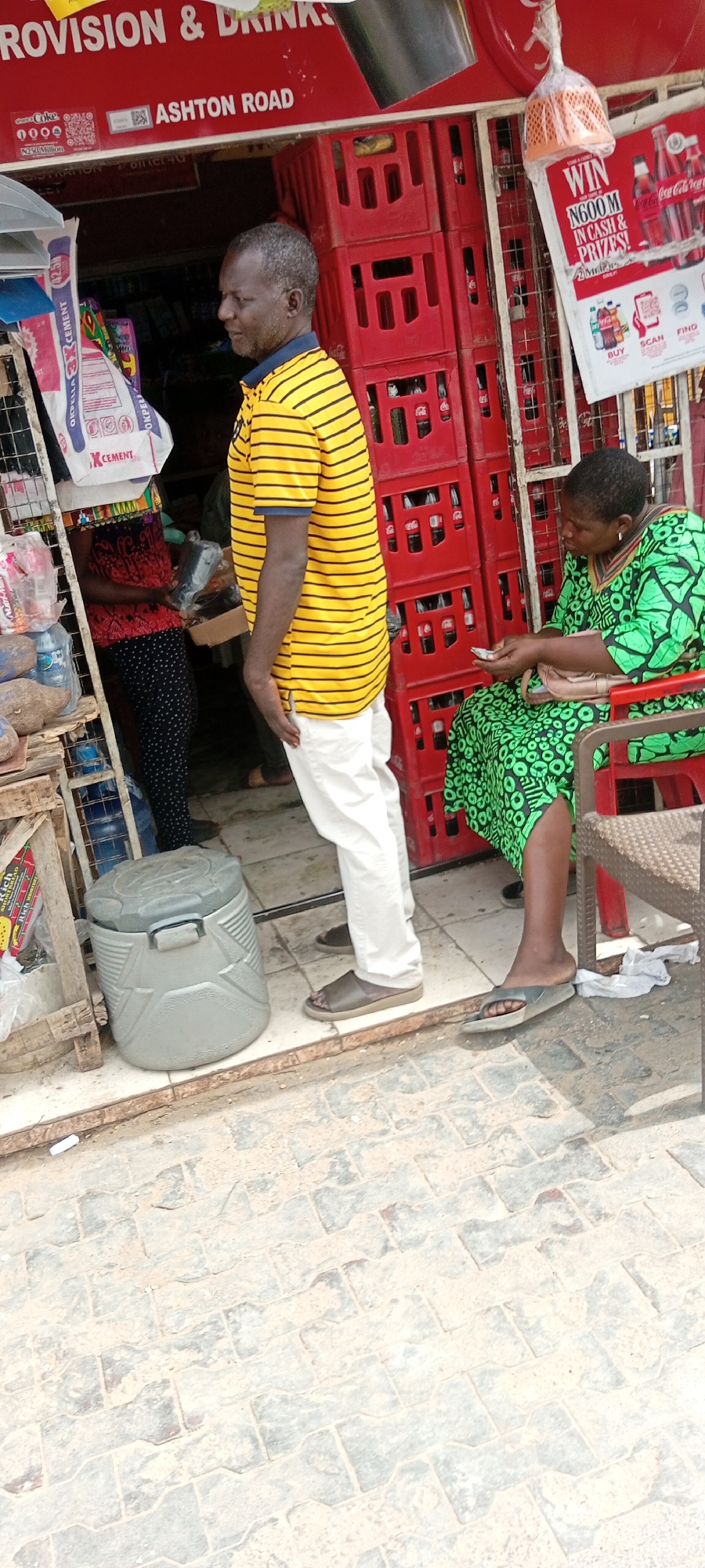The scene is set in front of a provision store located on Ashton Road in Kwachiri, Nigeria. A middle-aged man stands near the entrance of the store wearing a yellow and black striped shirt with white pants and brown sandals. He appears to be looking towards the store's interior, where a person wearing a red and black patterned outfit is being served. Beside the man is a grey cooler. To the right, an older woman wearing a green patterned dress sits on a red plastic chair, examining paper money. Behind her are stacks of red Coca-Cola crates. A sign advertising a Coca-Cola promotional giveaway hangs on the store's facade. The ground in front of the store is paved with grey interlocking bricks. The storefront has a red sign above it, reading "PROVISION & DRINKS" and "ASHTON ROAD". Bags of cement are visible inside the store, partially obscuring the view of the person being served.