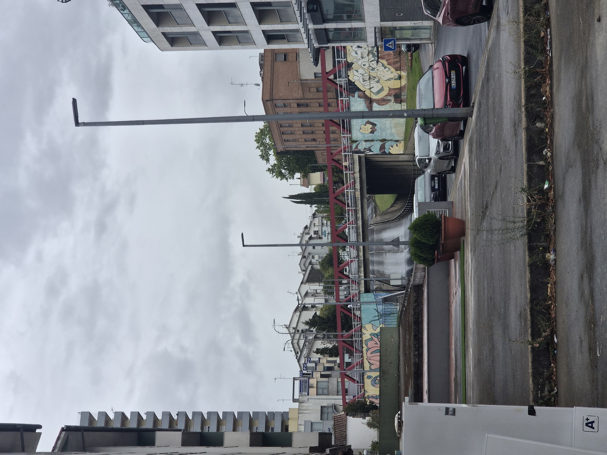 The image depicts an overcast day in Bragança, Portugal, likely on a street with a bridge crossing over. In the foreground, a wet asphalt road is visible, with several parked cars. A red sedan with license plate "63 ZR 40" is prominent, parked on the right side of the road. To its left, a silver hatchback and a dark gray sedan are also parked. A green trash bin is positioned between the red sedan and the silver hatchback. In the background, a pedestrian bridge with a red railing spans over a road or pathway. The bridge is adorned with graffiti art. Buildings, including a tall apartment complex on the left and modern multi-story structures on the right, line the street. A lamppost stands near the right edge of the image. The scene appears to be a typical urban street environment, possibly in a residential or commercial area.