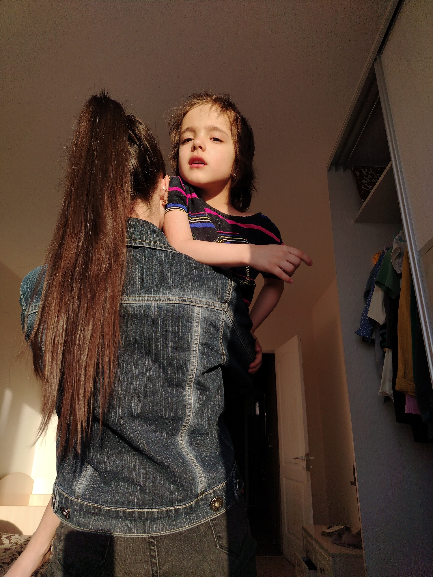 A woman stands with a child in her arms in an indoor setting. The woman is seen from the back, with long brown hair pulled into a high ponytail. She wears a denim jacket over dark clothing. The child, held in the woman's arms, looks towards the viewer and points with their right hand. They wear a striped shirt. The room includes a closet with clothes hanging inside, a white door and a light-colored wall. The objects and setting provide insight, but cannot confirm the specified location in Noviy, Russia.