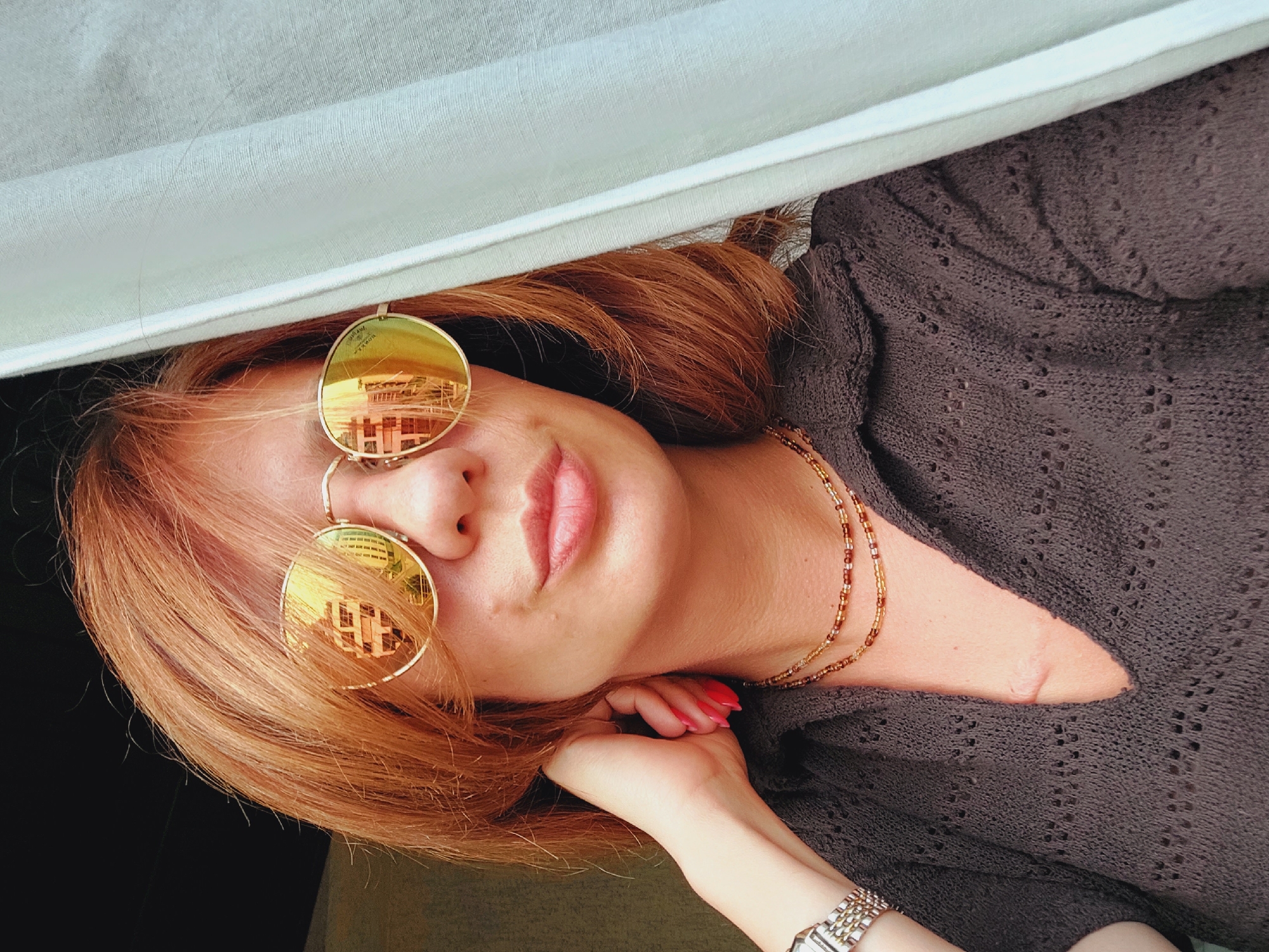 This is a vertically oriented, close-up, selfie-style photograph of a woman. She has reddish-brown hair and is wearing gold-rimmed, reflective sunglasses that show a reflection of a building, possibly in Sofia, Bulgaria. Her lips are slightly parted, and she has pink nail polish on her fingers. She is wearing a dark, knitted top with a diamond pattern and a beaded necklace made of small gold and brown beads. Her right hand is resting near her chin, and on her left wrist, she is wearing a silver watch with a rectangular face. The background consists of a light blue fabric draped from above and a dark surface below. The lighting suggests it could be daytime, and the overall mood is casual and relaxed.