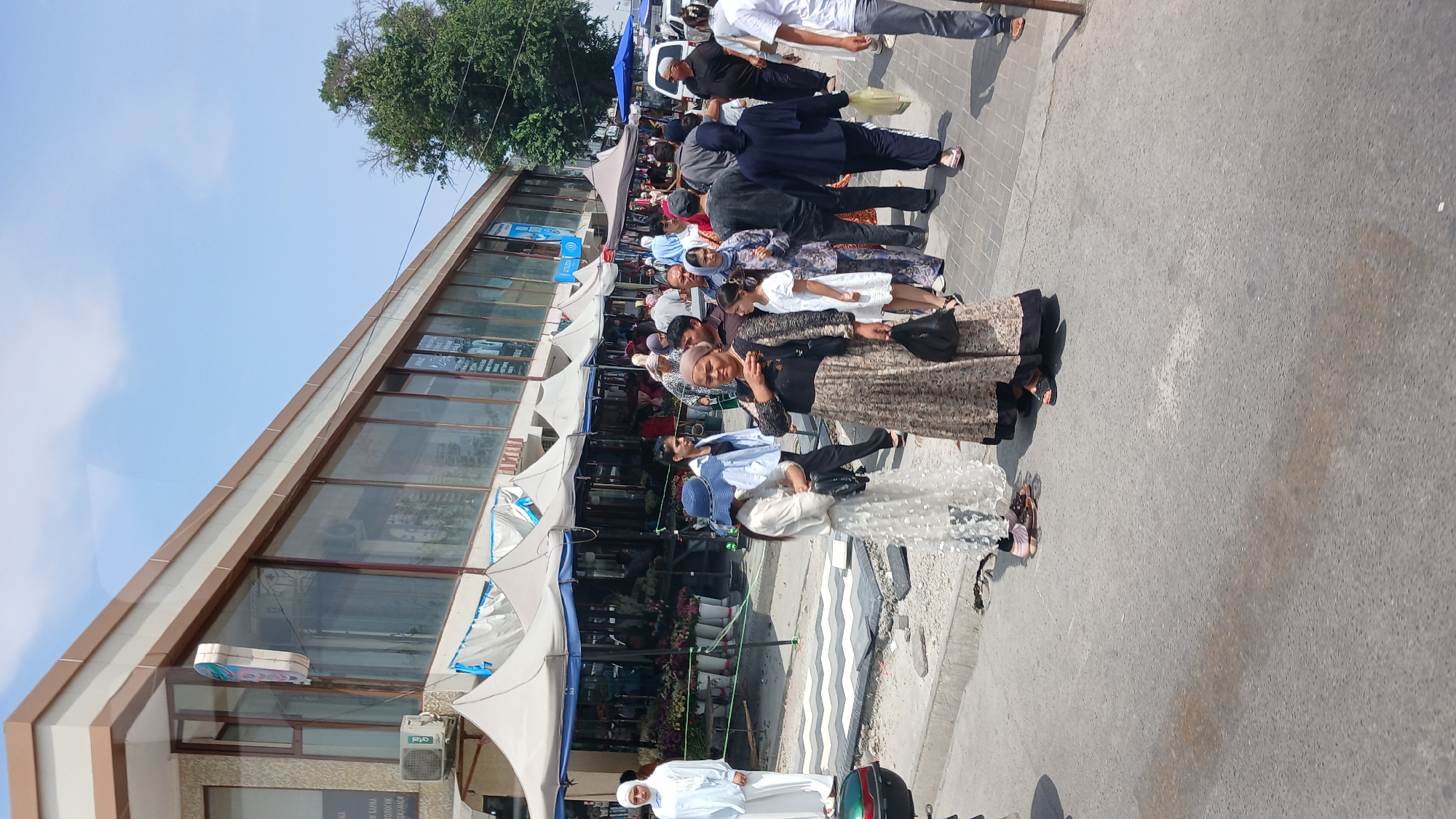 In Fergana City, Uzbekistan, a bustling street scene unfolds in front of a row of market stalls and shops. The architecture of the shops features large glass windows and a beige and brown facade, with white awnings extending over the stalls. A diverse group of people, dressed in a mix of traditional and modern attire, are walking and shopping.
Several individuals stand out: a woman in a light blue hijab and white abaya on the left, a young girl in a white dress with a blue hat, a woman in a patterned dress carrying a black purse, and a woman in a dark blue hijab and trousers. Other shoppers are visible throughout the scene, some carrying bags.
The ground is paved with asphalt and a patterned walkway. The stalls are filled with an abundance of flowers, suggesting a flower market. The weather appears to be sunny, with a clear blue sky visible above. The scene captures the everyday life and commerce of this urban environment.