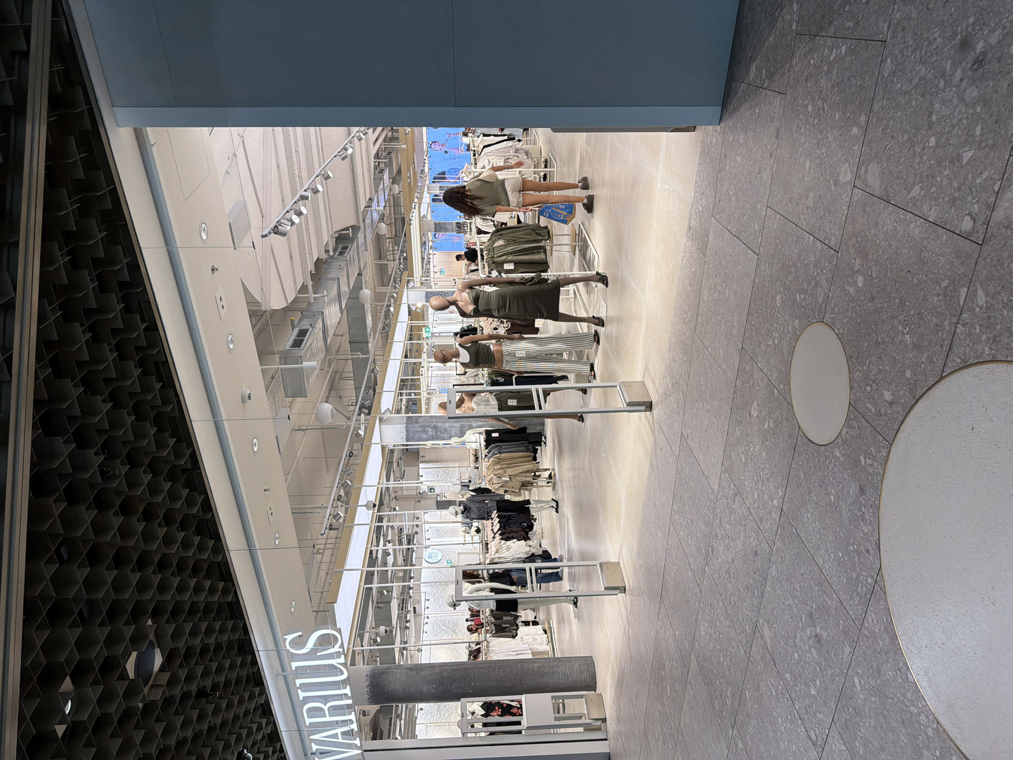This is a medium shot, taken at eye level, of the entrance to a clothing store. The store's name, "VARIUS", is visible in large white letters on the facade. The entrance is open and has a security gate system at the doorway. Inside, mannequins are displayed wearing various outfits, and racks of clothing are visible. The store has a modern interior with a tiled floor and a patterned ceiling. Several people are seen browsing or walking through the store. The exterior is a bright, neutral color. The overall impression is of a clean and well-lit retail environment.